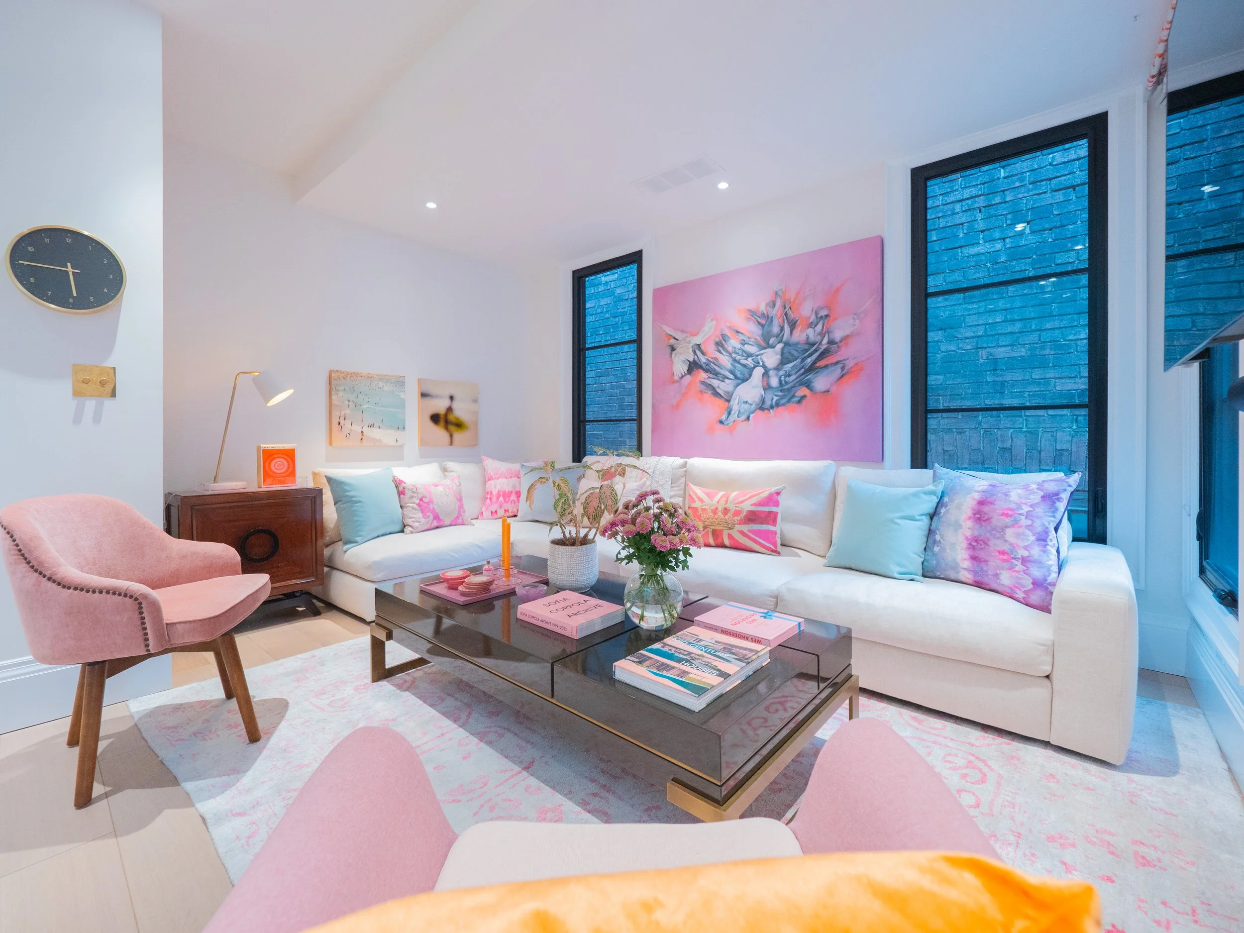 A cozy living room with white sectional sofa with pink and pastel pillows, a pink upholstered armchair, a coffee table with flowers and magazines, wall art with pink tones, and large windows with blue bricks visible outside.
