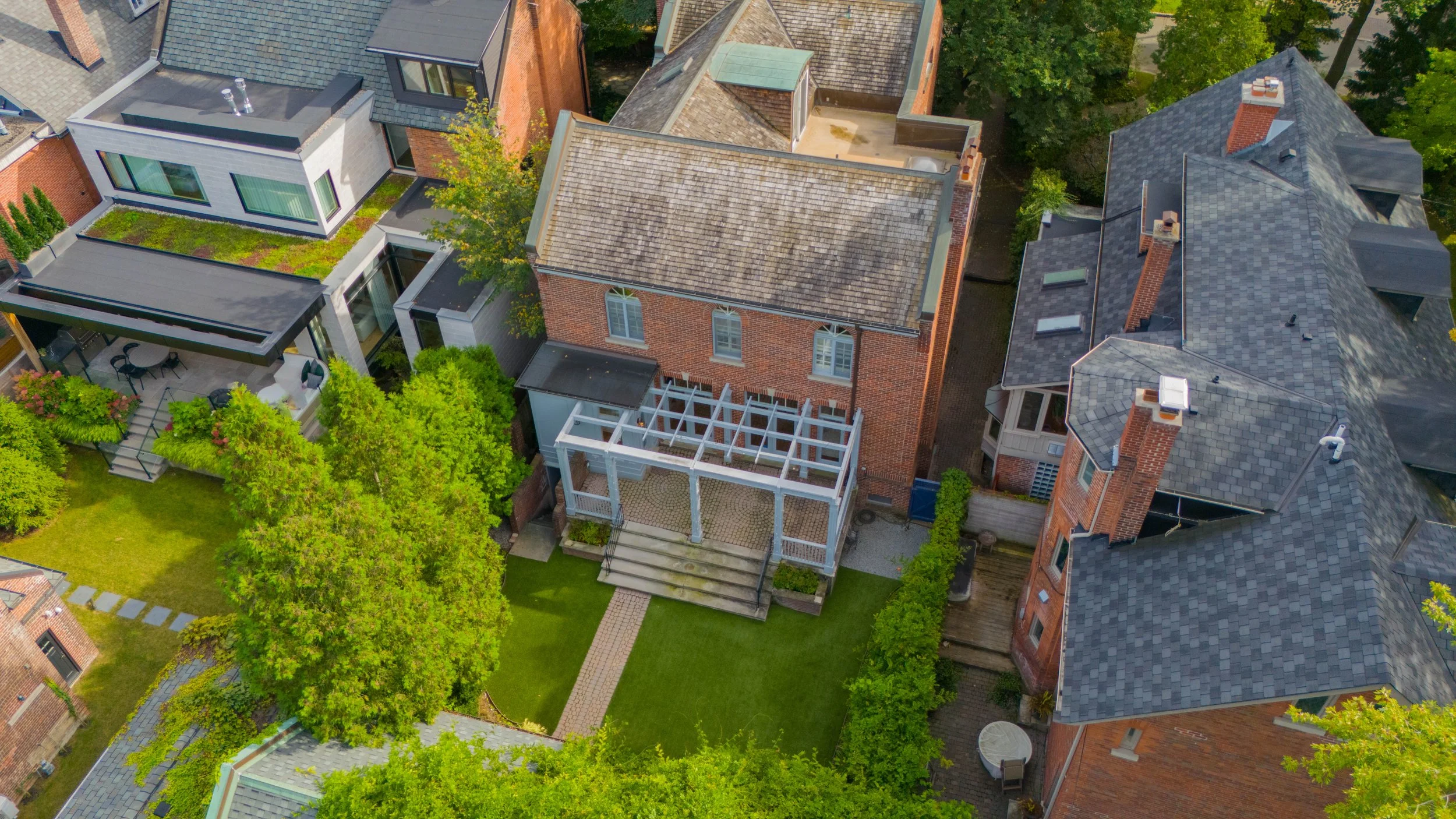Aerial view of a Toronto neighborhood with multiple houses and lush green trees, focusing on a brick house with a wooden deck and green lawn.