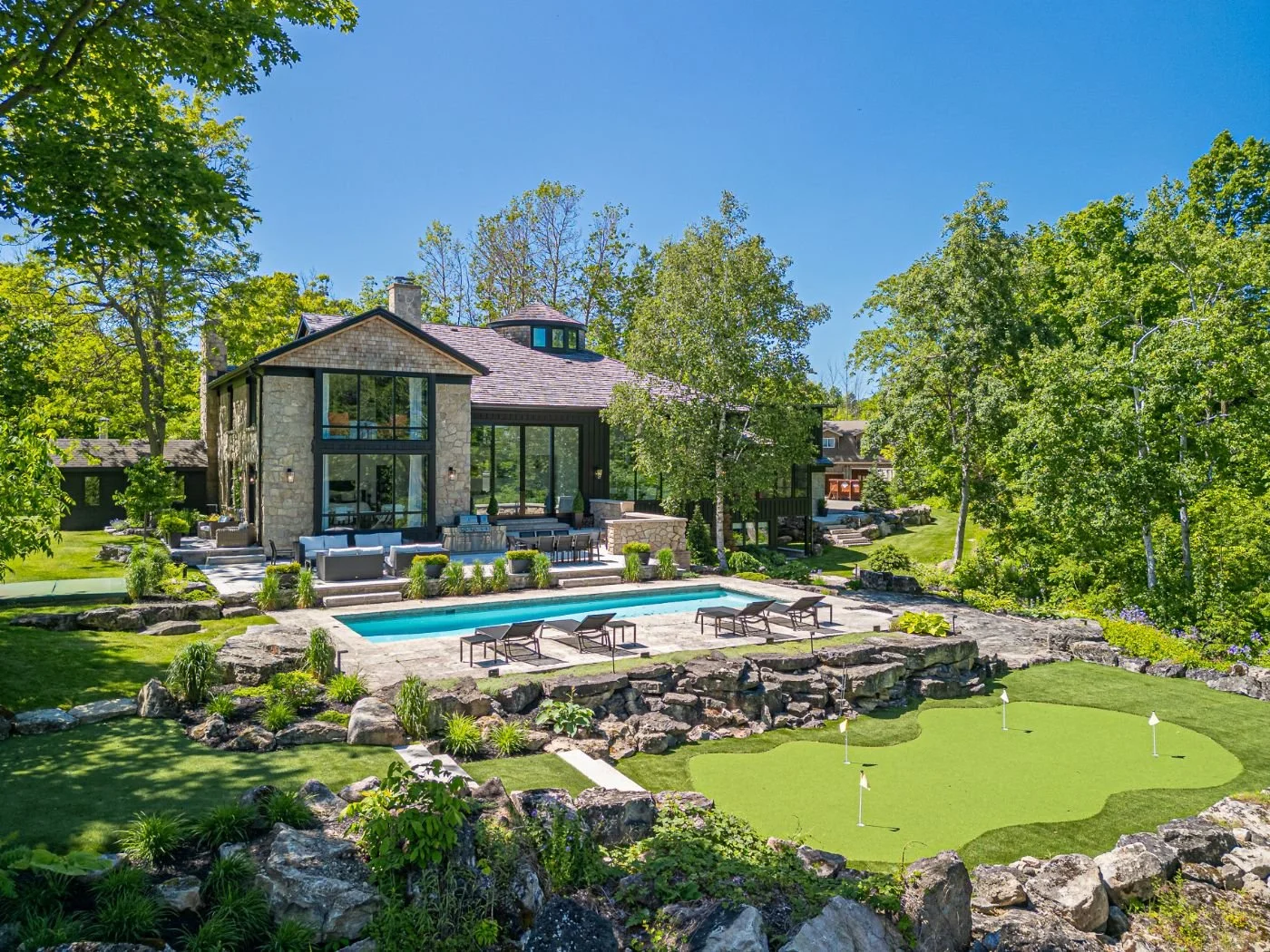 Luxury house with large windows, pool, and a lawn with a putting green, surrounded by trees on a sunny day.