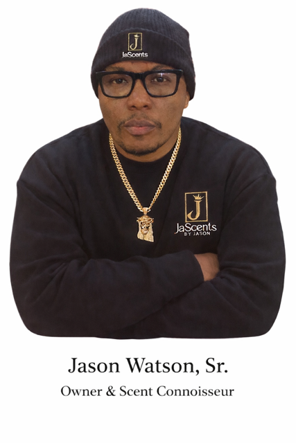 A man wearing a black beanie, glasses, and a black sweatshirt with a JeSi scents logo, accessorized with a gold chain and pendant, standing with arms crossed.
