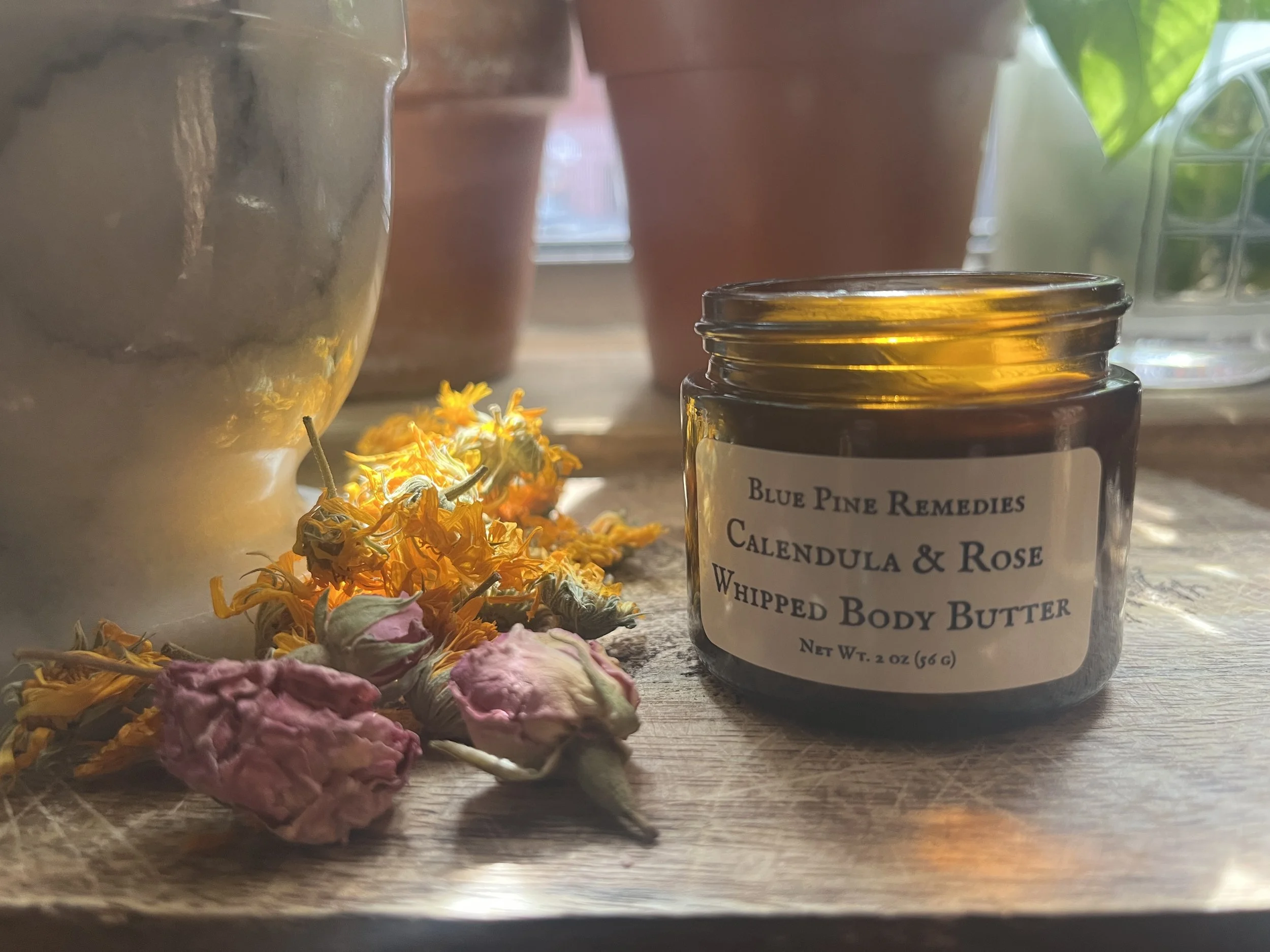 A small jar of Blue Pine Remedies Calendula & Rose Whipped Body Butter on a wooden surface, surrounded by dried flowers, with potted plants and a window in the background.