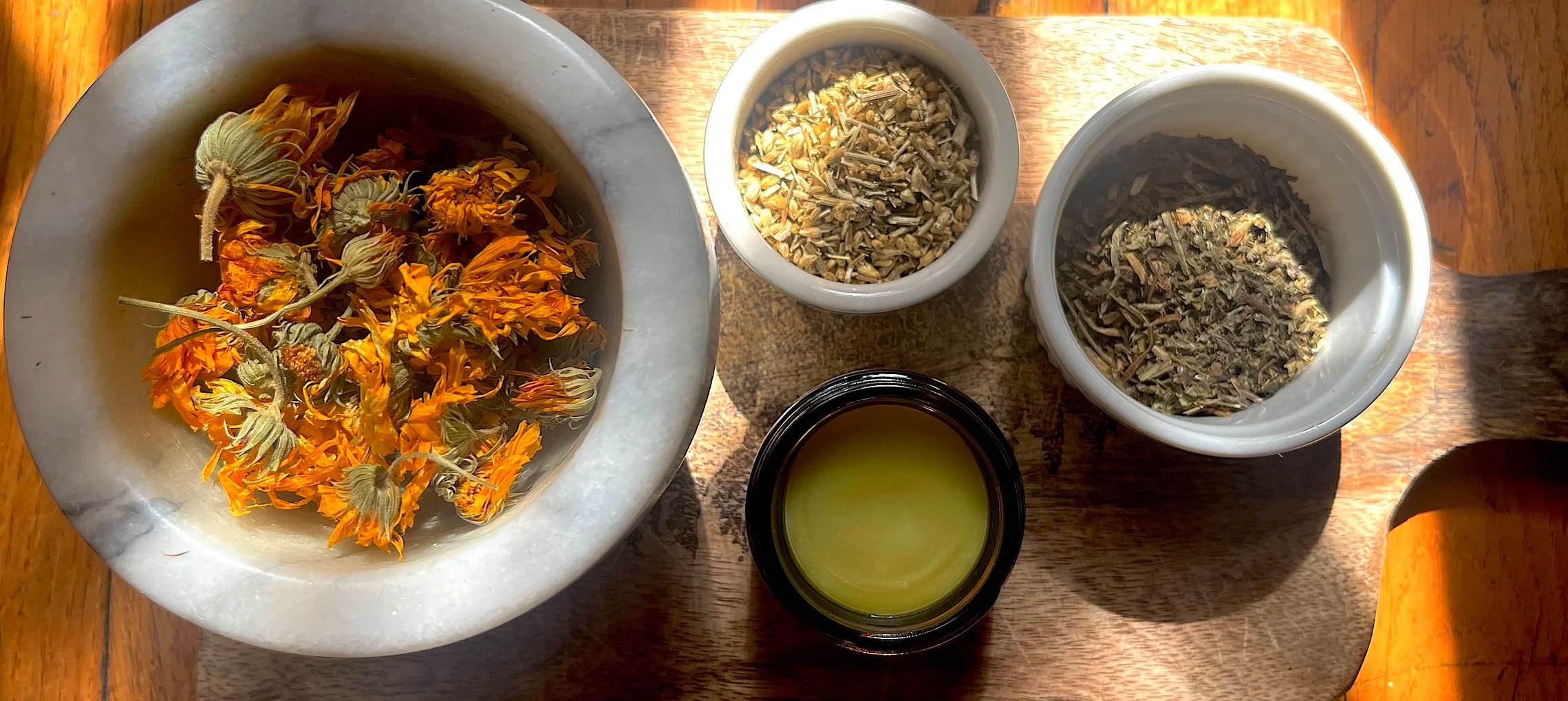 A wooden table holding a marble mortar with dried orange flowers, two small white bowls with dried herbs, a small black container with yellowish green oil or syrup, and a larger white bowl with dried herbs and flowers.