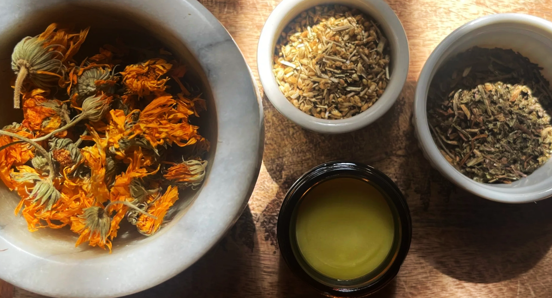 Dried flower petals and herbs in bowls and a small jar of green oil or liquid on a wooden surface.