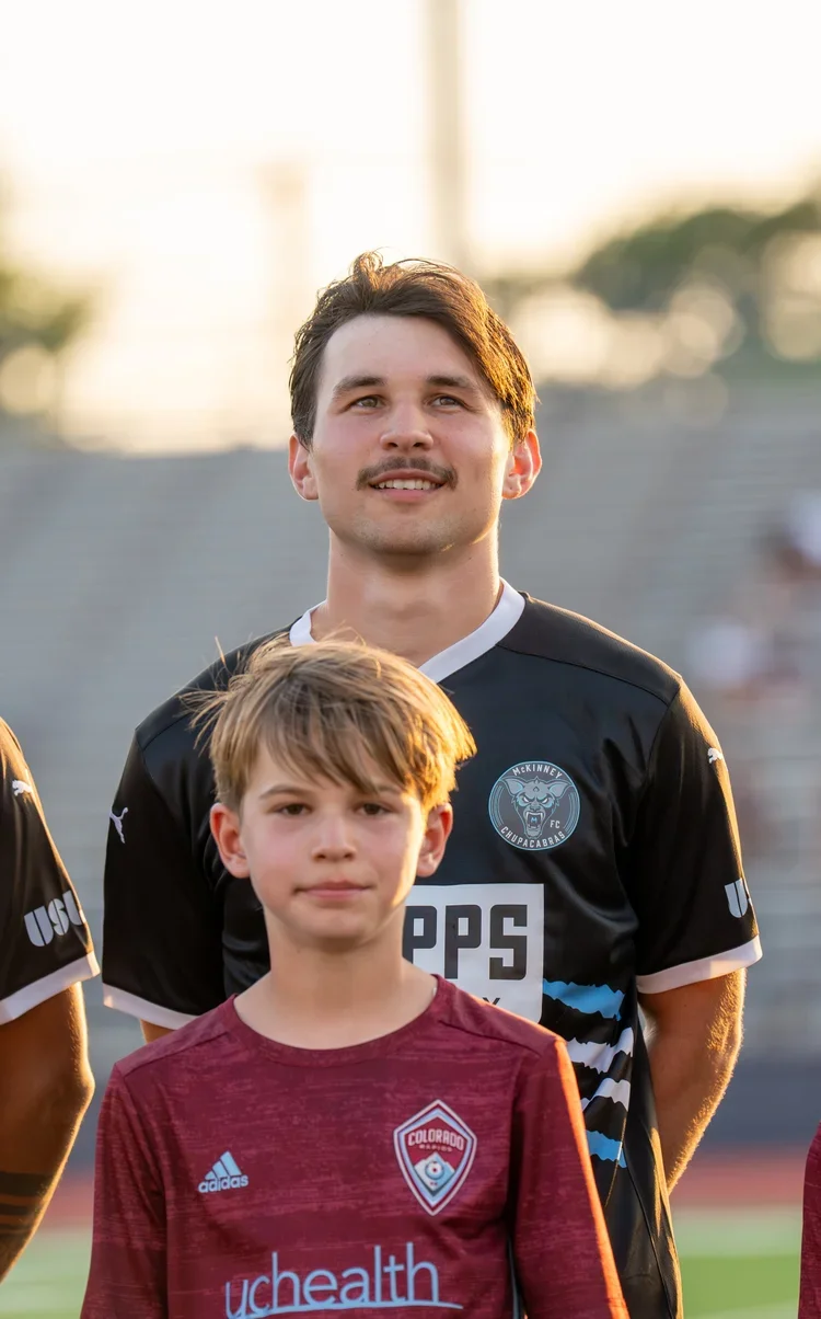 A young man and a boy standing on a football field during sunset, both wearing sports jerseys.