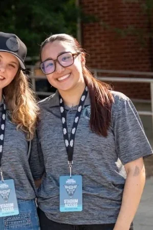 Two young women smiling outdoors, wearing gray shirts, with stadium access passes around their necks.
