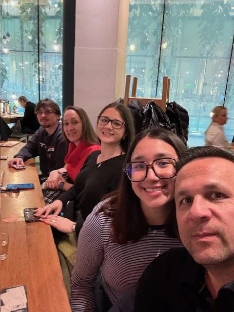 Five people sitting at a restaurant table, smiling, with large windows behind them.