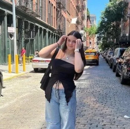 A young woman standing on a city street with parked cars, buildings, and trees in the background. She is wearing a black tank top, light blue jeans, and has a black bag over her shoulder.