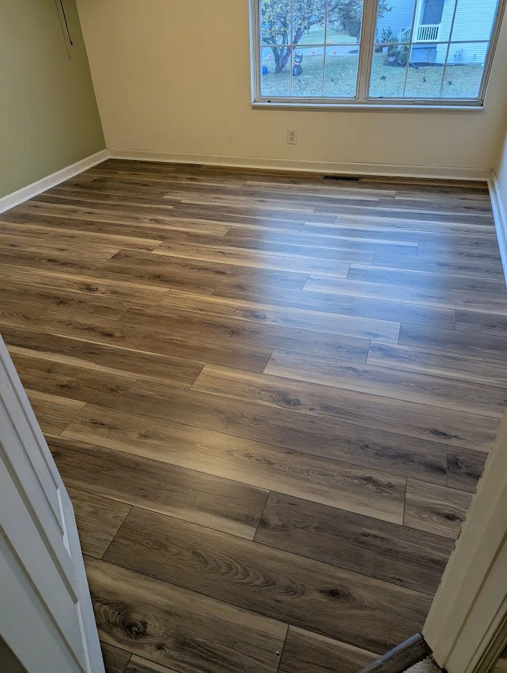 Empty room with wood-like flooring, a large window showing a yard with a tree and some outdoor objects, cream-colored walls, and white baseboards.
