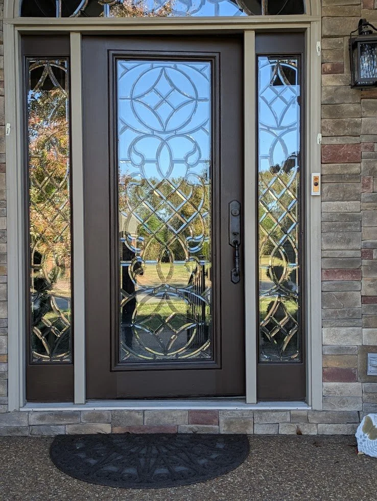 A front door with decorative glass panels on each side, featuring geometric and floral patterns, with a black doormat in front and a brick exterior wall.