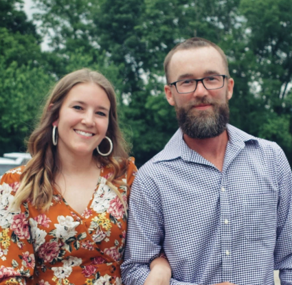 A smiling woman and a man with a beard and glasses standing outdoors in front of trees, with the woman wearing a floral dress and the man wearing a checkered shirt.