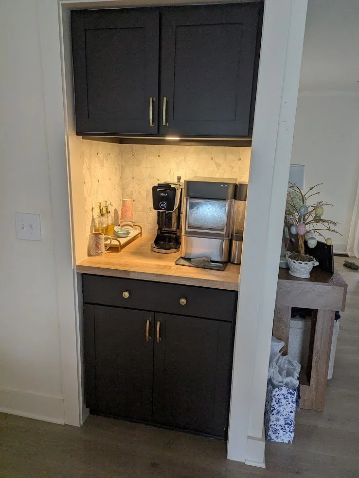 A small coffee station with a coffee maker and a water dispenser on a wooden countertop, with dark cabinets above and below.