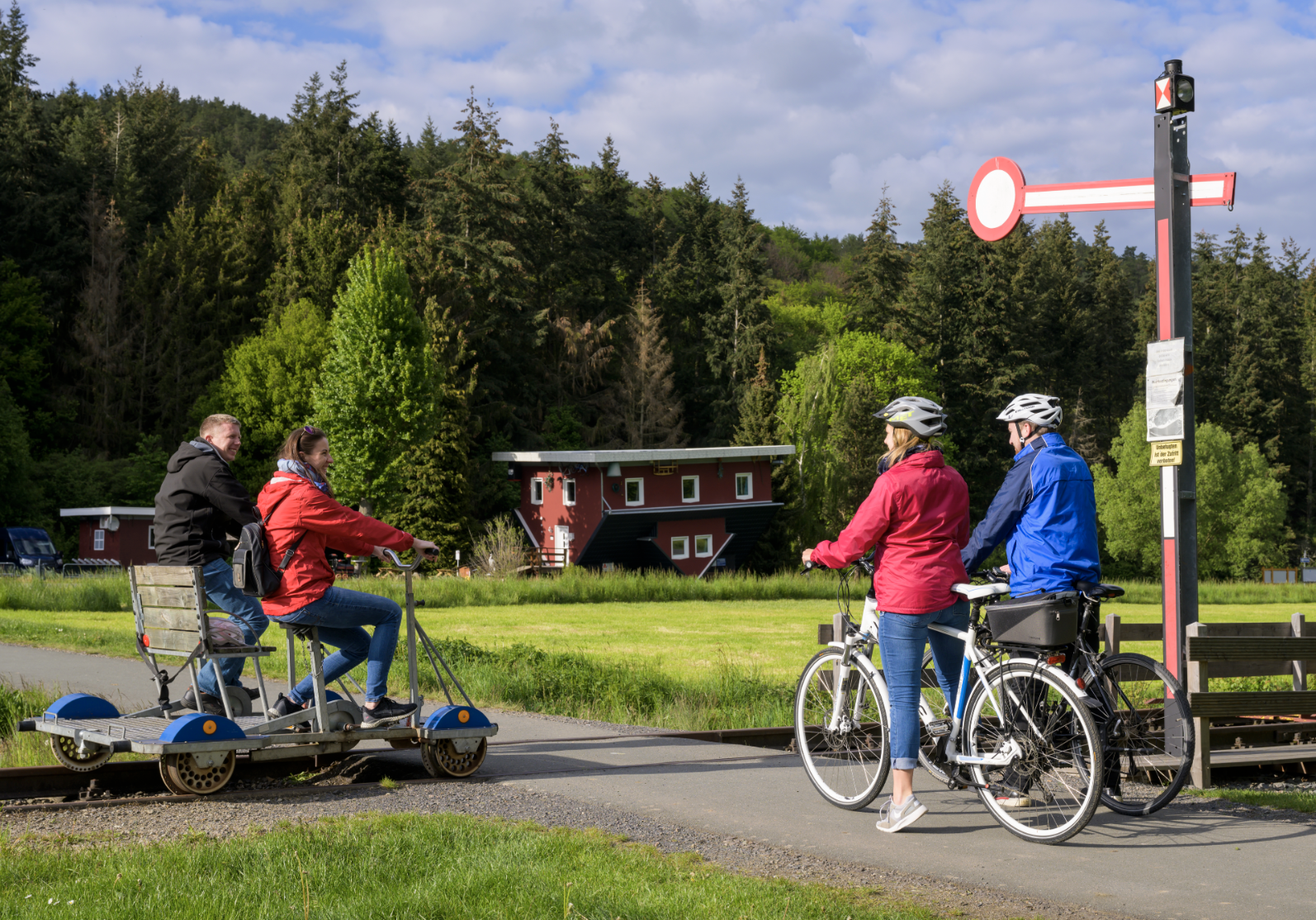 Vier Personen beim Radfahren und Tretroller fahren in einer grünen, ländlichen Gegend mit Bäumen und modernen Gebäuden im Hintergrund.