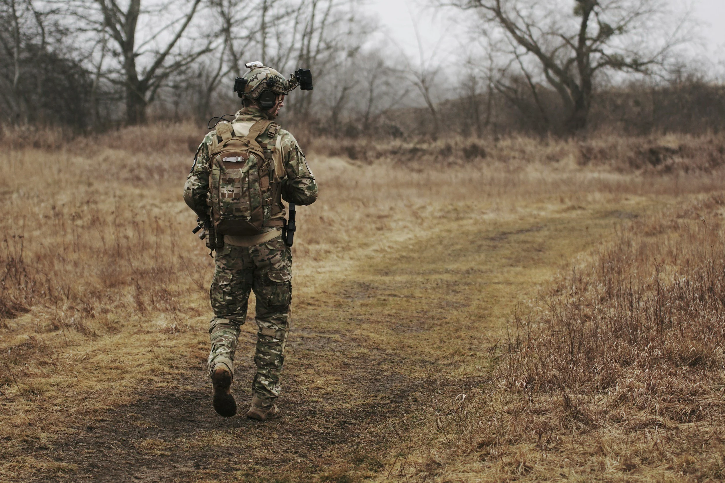 A soldier in camouflage gear walking on a dirt trail through a barren field with leafless trees in the background.