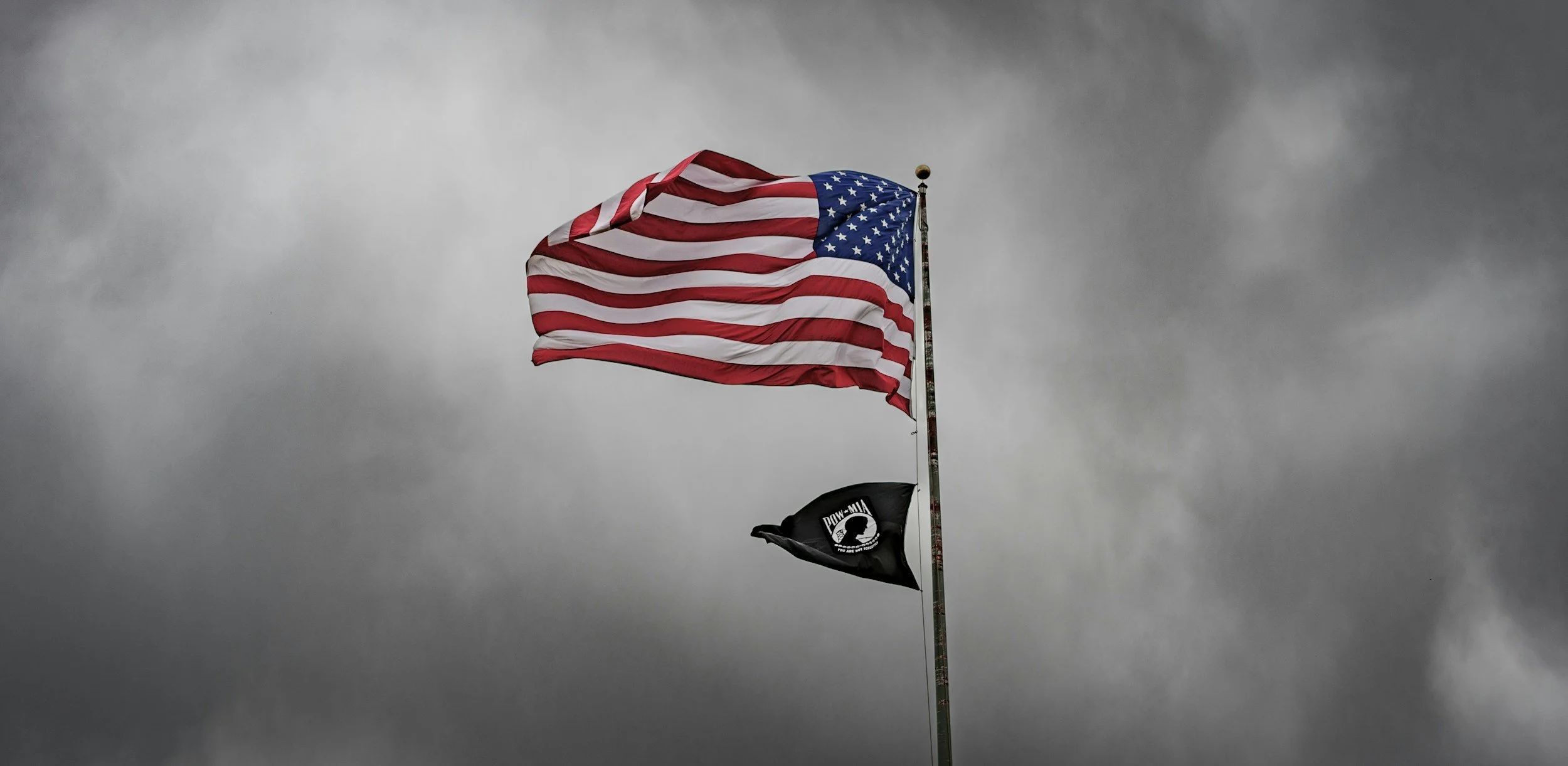 American flag and black POW-MIA flag flying against a cloudy, overcast sky.