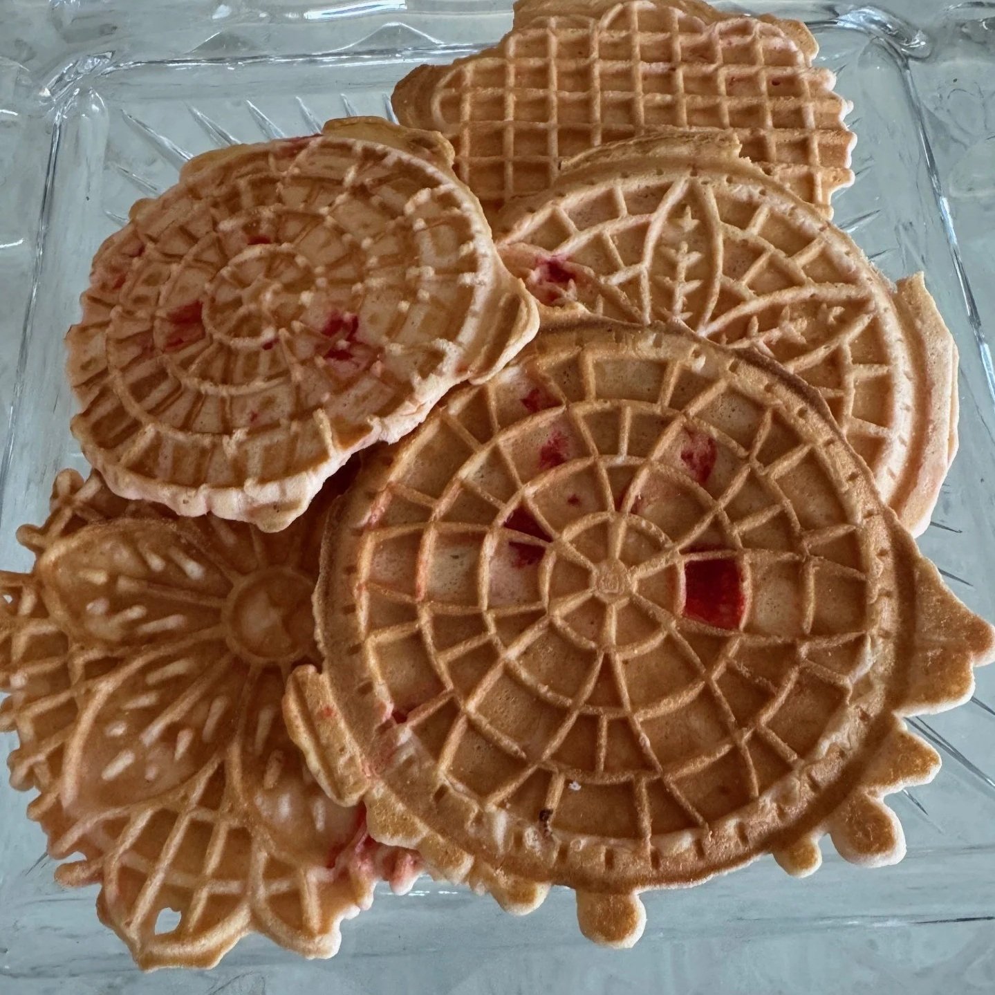 Several round waffle cookies with a crisscross pattern, some with red fruit filling, arranged on a clear glass tray.