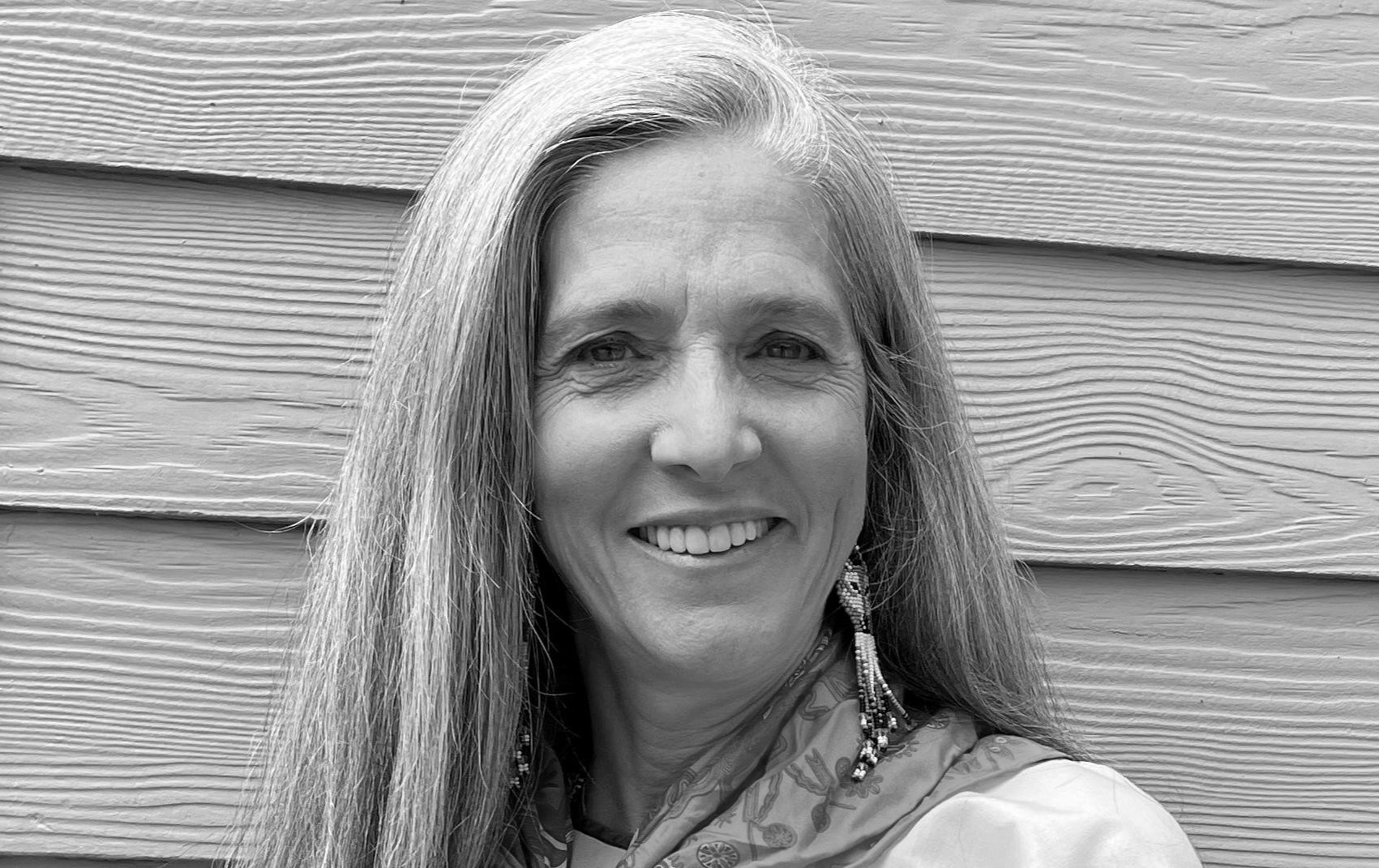 Retrato en blanco y negro de mujer sonriendo, con cabello largo y ondulado, usando pendientes largos y bufanda con patrón, frente a fondo de tablones de madera horizontal.