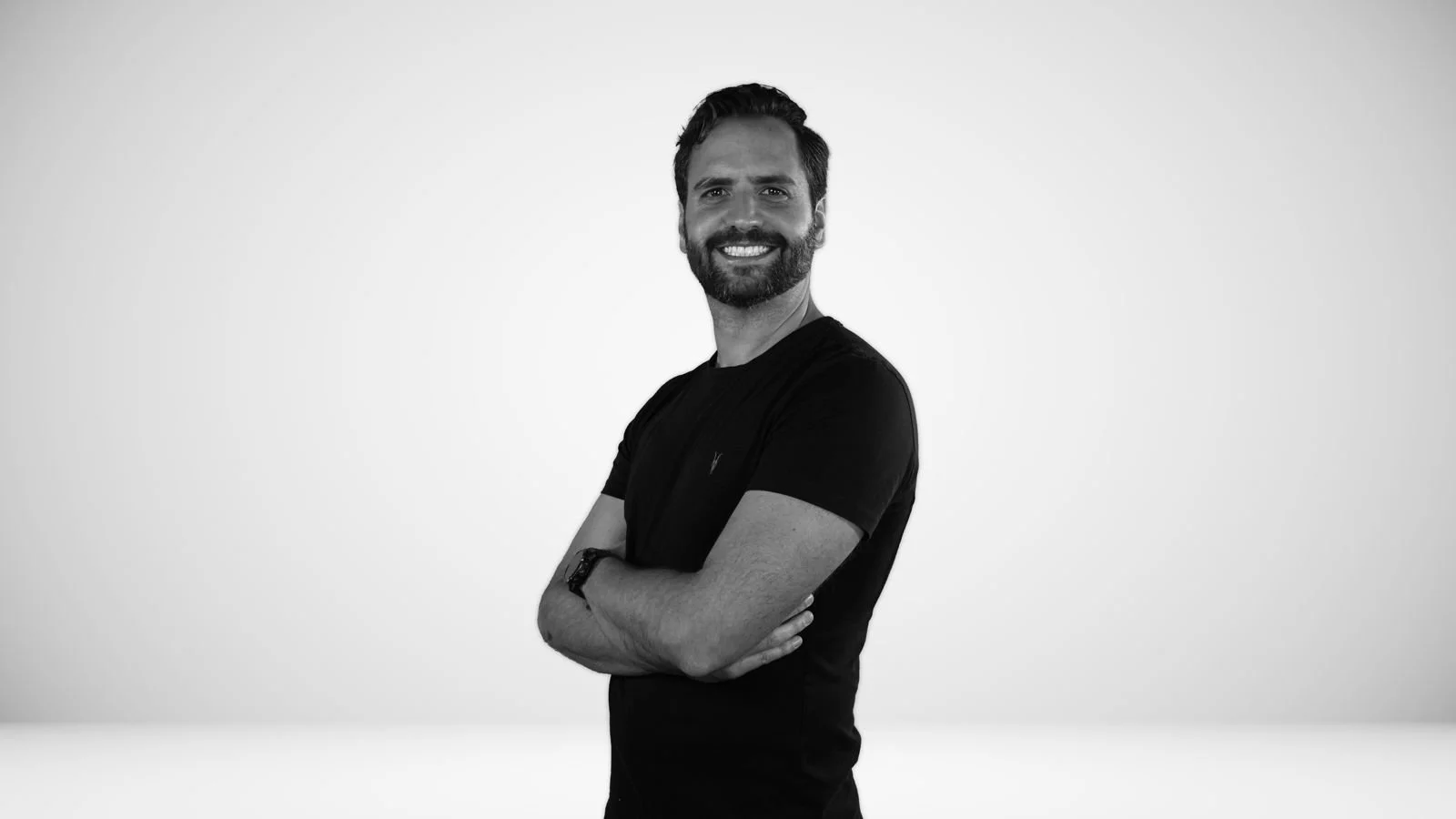 Hombre con barba y cabello oscuro, sonriendo, usando ropa negra con los brazos cruzados, en un fondo gris liso.