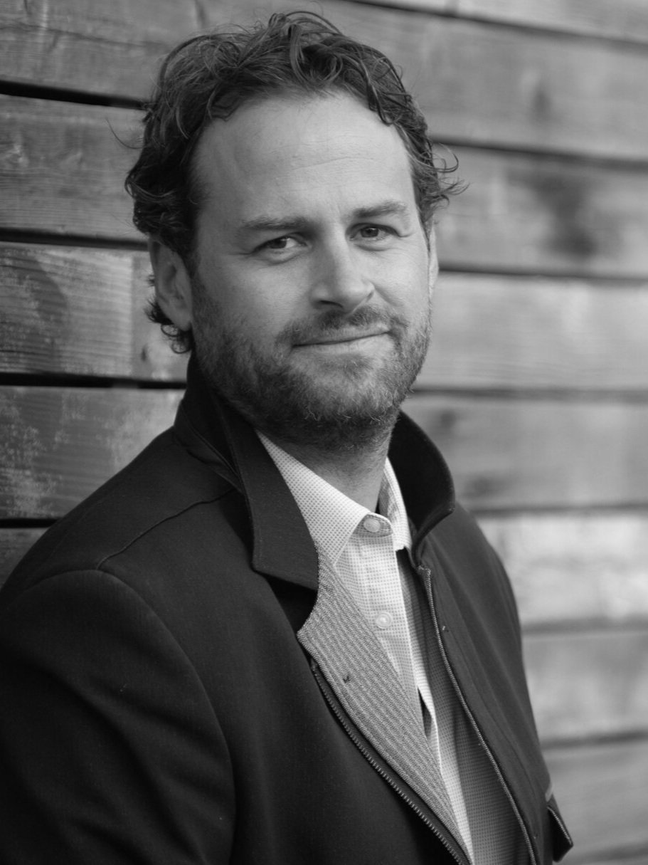 Retrato en blanco y negro de un hombre con barba, cabello rizado y expresión amable, vestido con chaqueta y camisa, posando junto a una pared de madera.