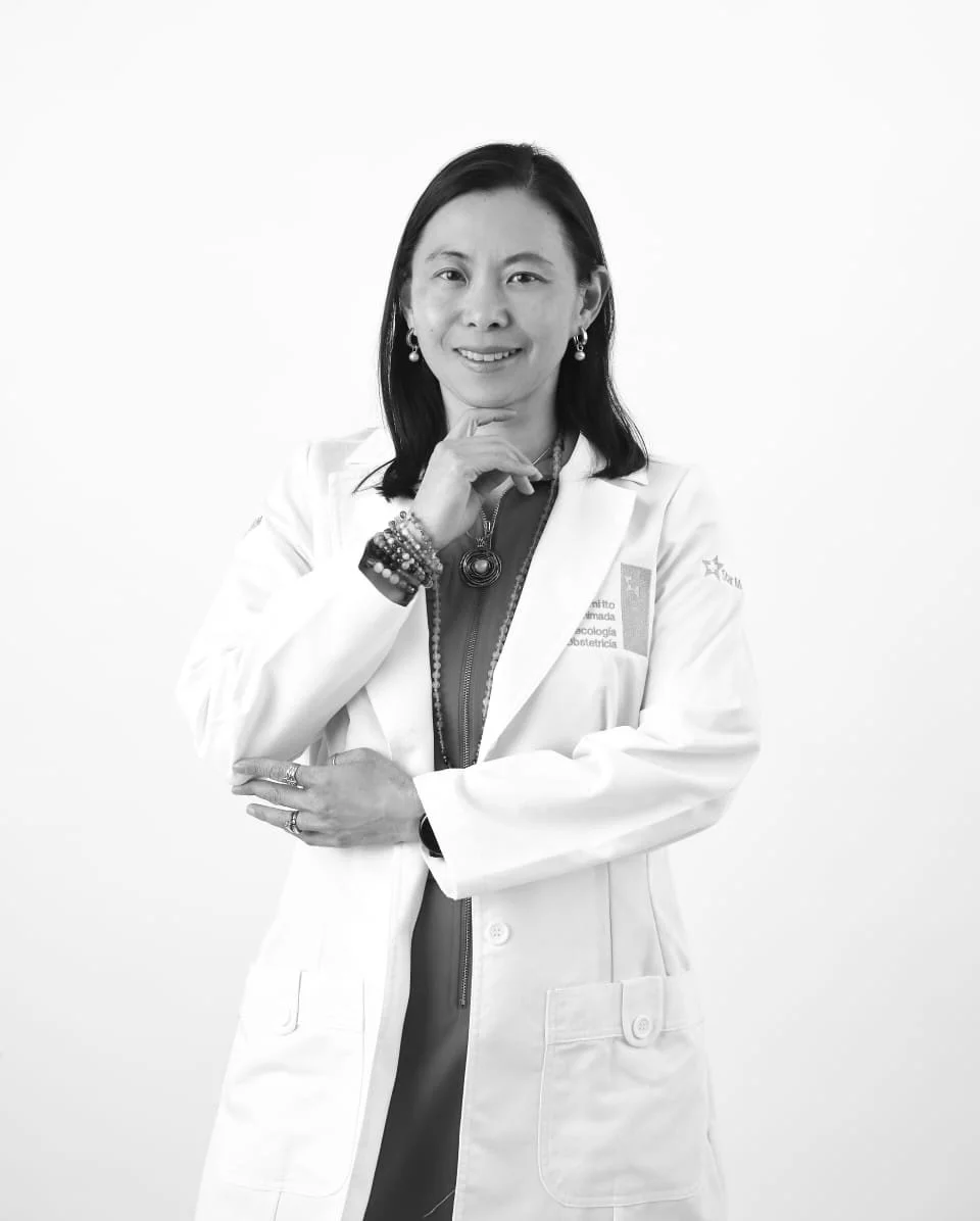 Mujer profesional sonriendo, con bata blanca, de cabello oscuro, usando accesorios y en un fondo blanco.