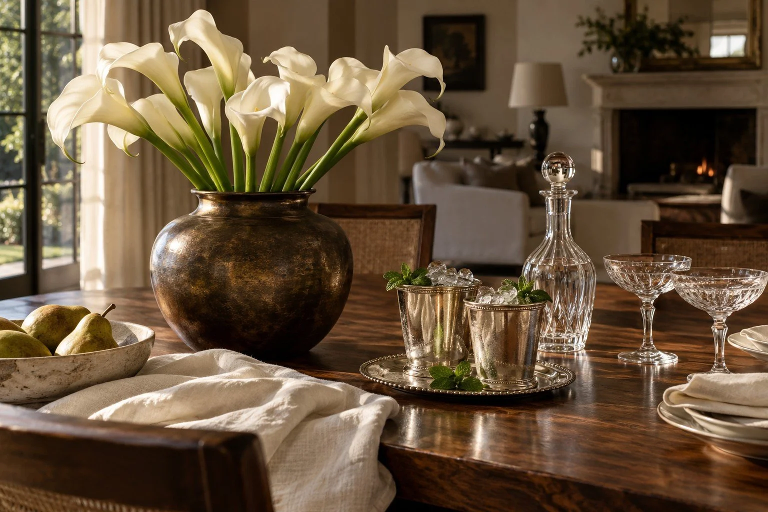 A dining table with a large vase of white calla lilies, a bowl of pears, glassware, and a decanter, in a well-lit room with a fireplace and living area in the background.