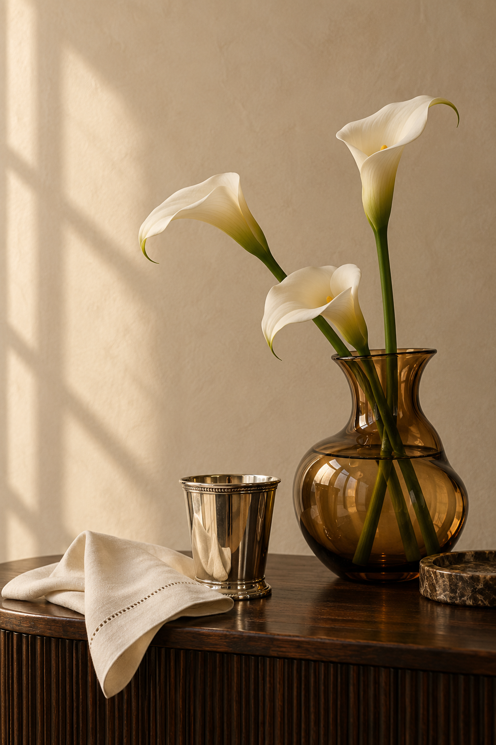 Three white calla lilies in a brown glass vase on a wooden table, with a small silver cup, a beige cloth, and a marble coaster.
