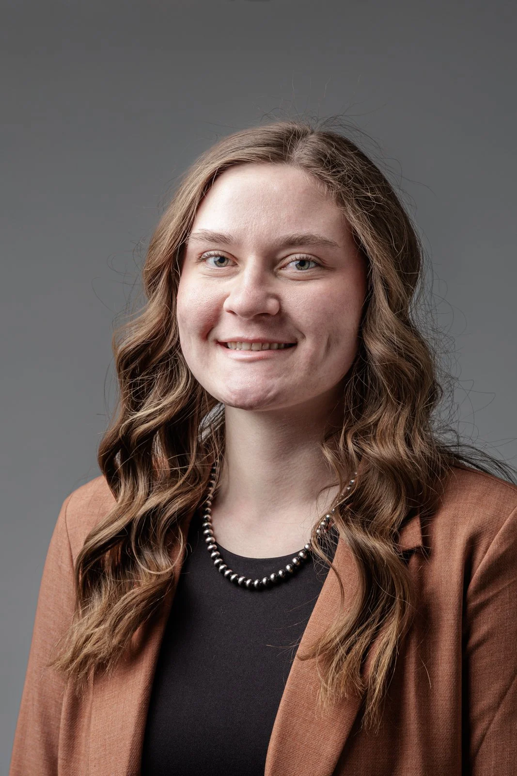 Portrait of a young woman with long curly brown hair, light skin, wearing a pearl necklace, black top, and brown blazer, smiling against a gray background.