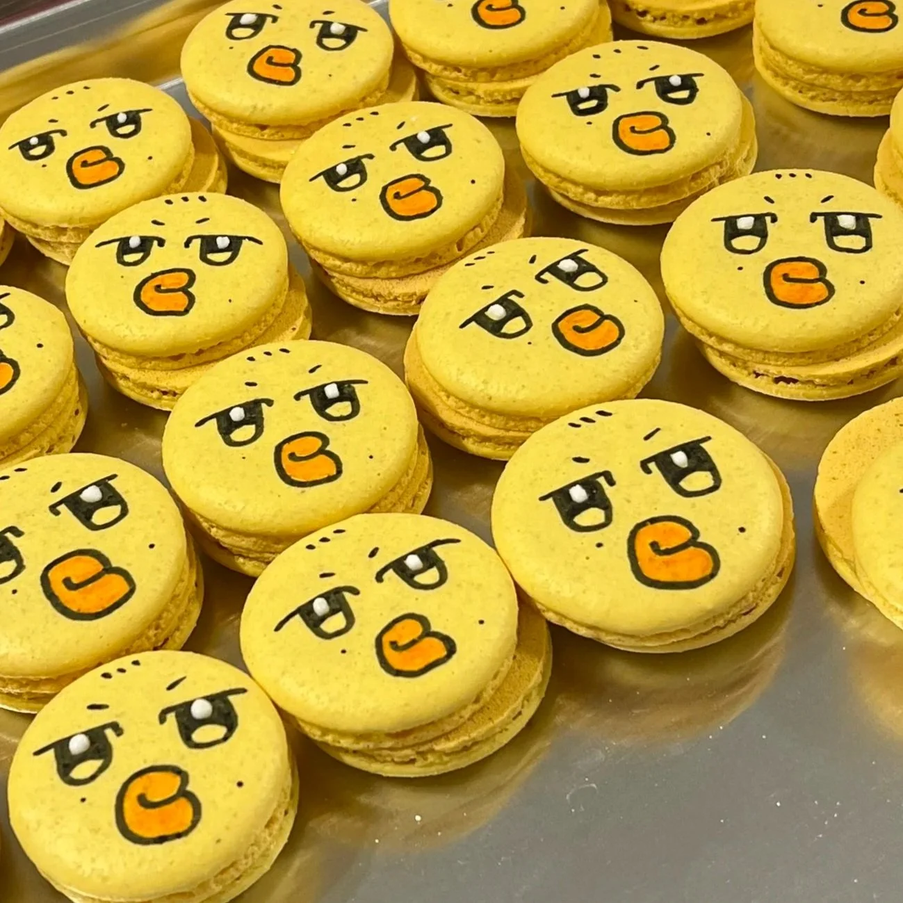 Multiple yellow macarons decorated with angry face emojis drawn with black and orange icing.