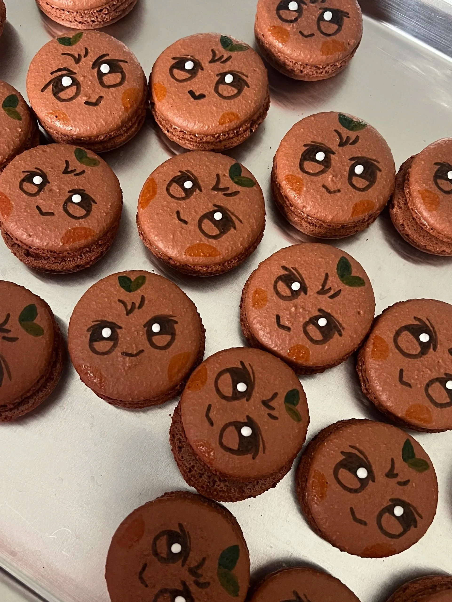 Close-up of multiple macarons with faces drawn on top, showing different facial expressions, on a tray.