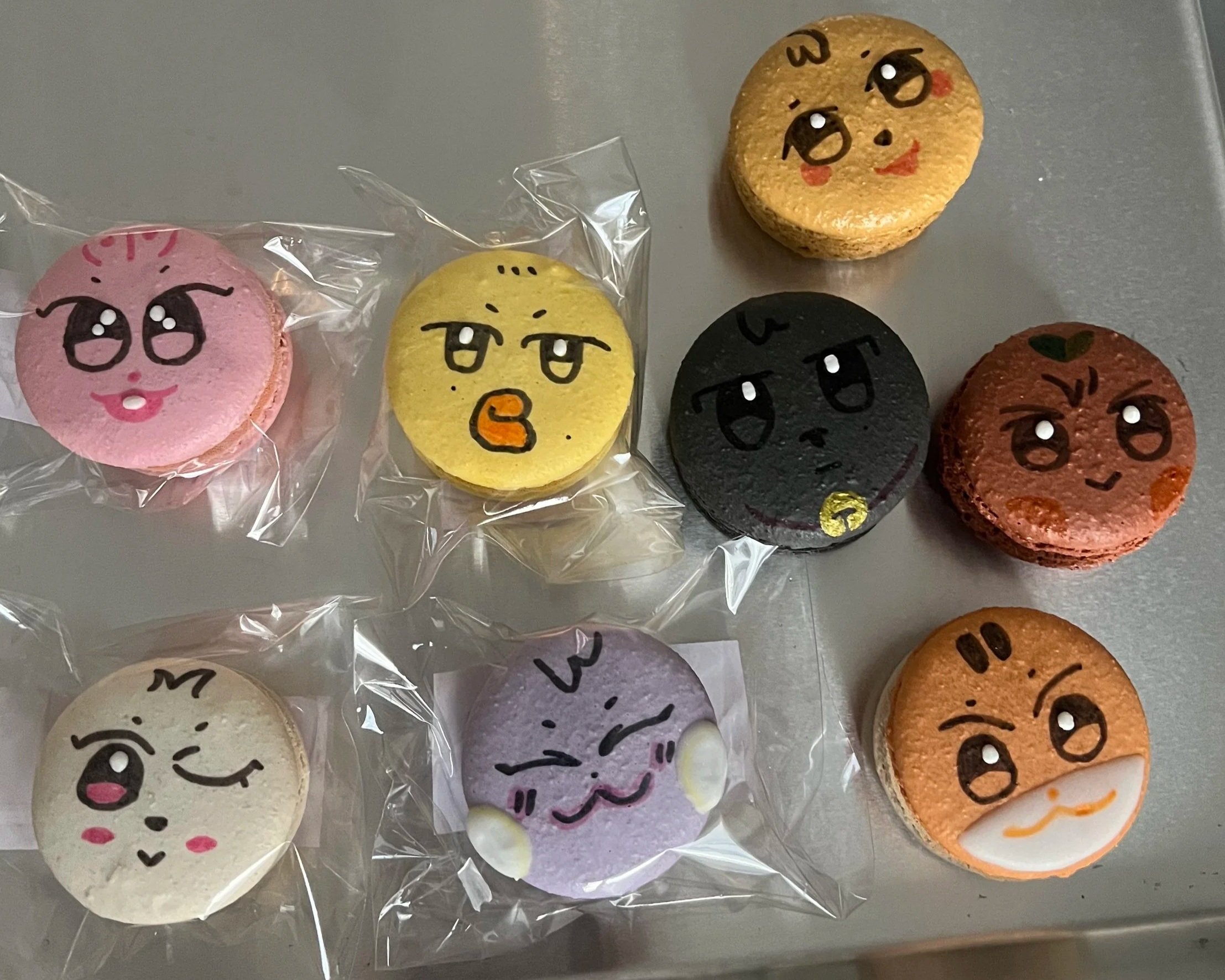 Decorative macarons with cartoon faces, some in plastic wrapping, in various colors including pink, yellow, black, white, and orange, arranged on a light gray surface.