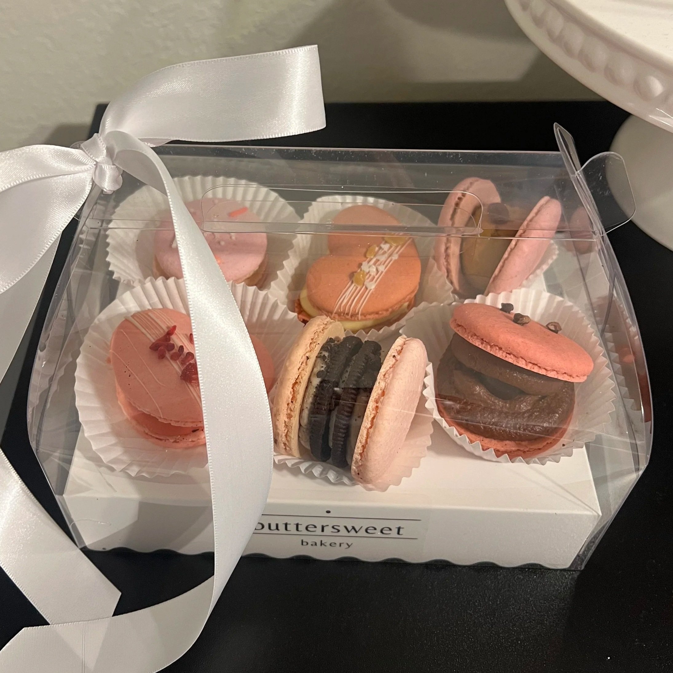 Clear plastic box with assorted macarons inside, tied with a white satin ribbon. The box is on a black tray with a white paper underneath that reads 'buttersweet bakery.'