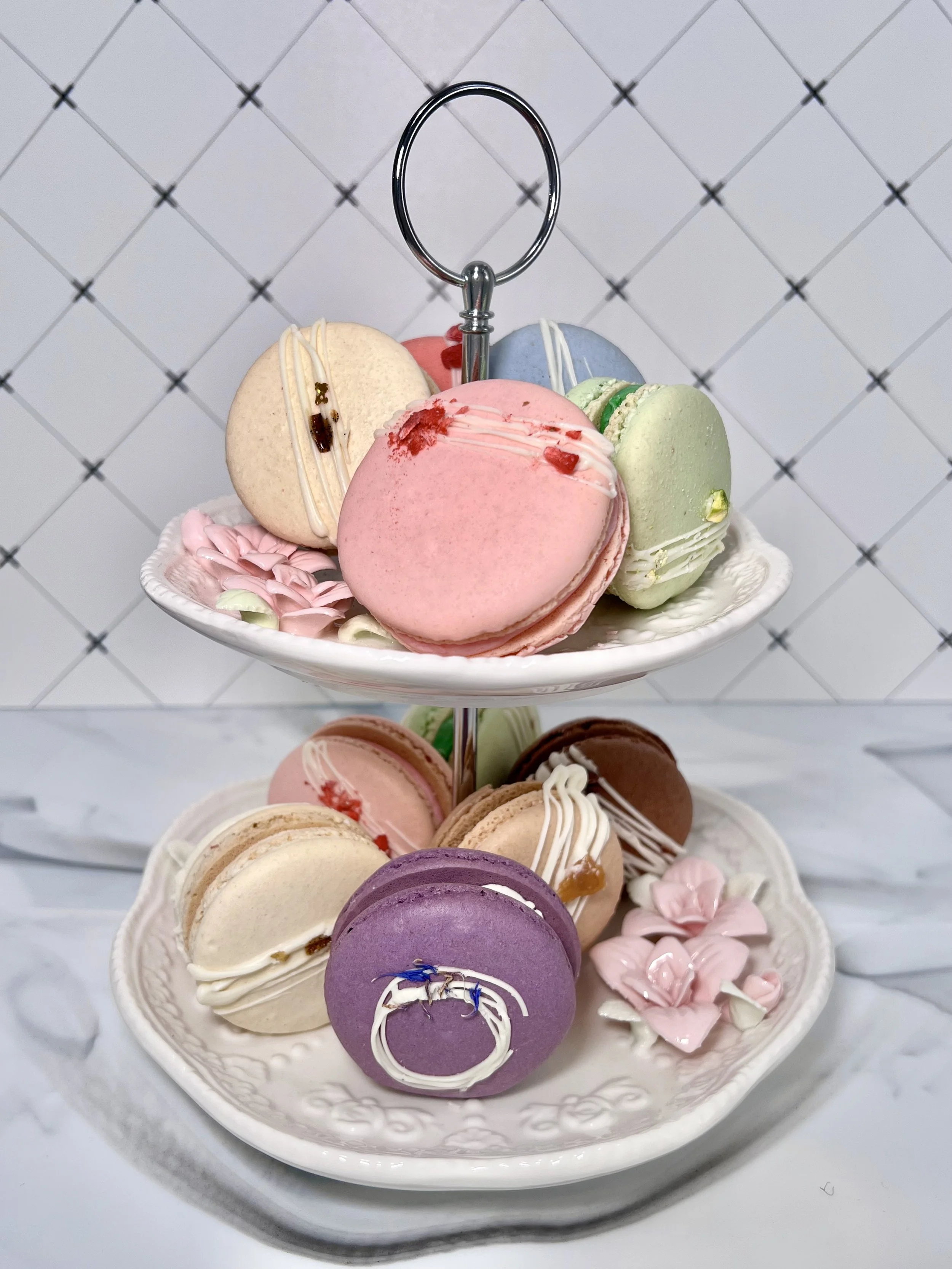 Decorative plate of colorful macarons on a tiered serving tray against a white tiled background.