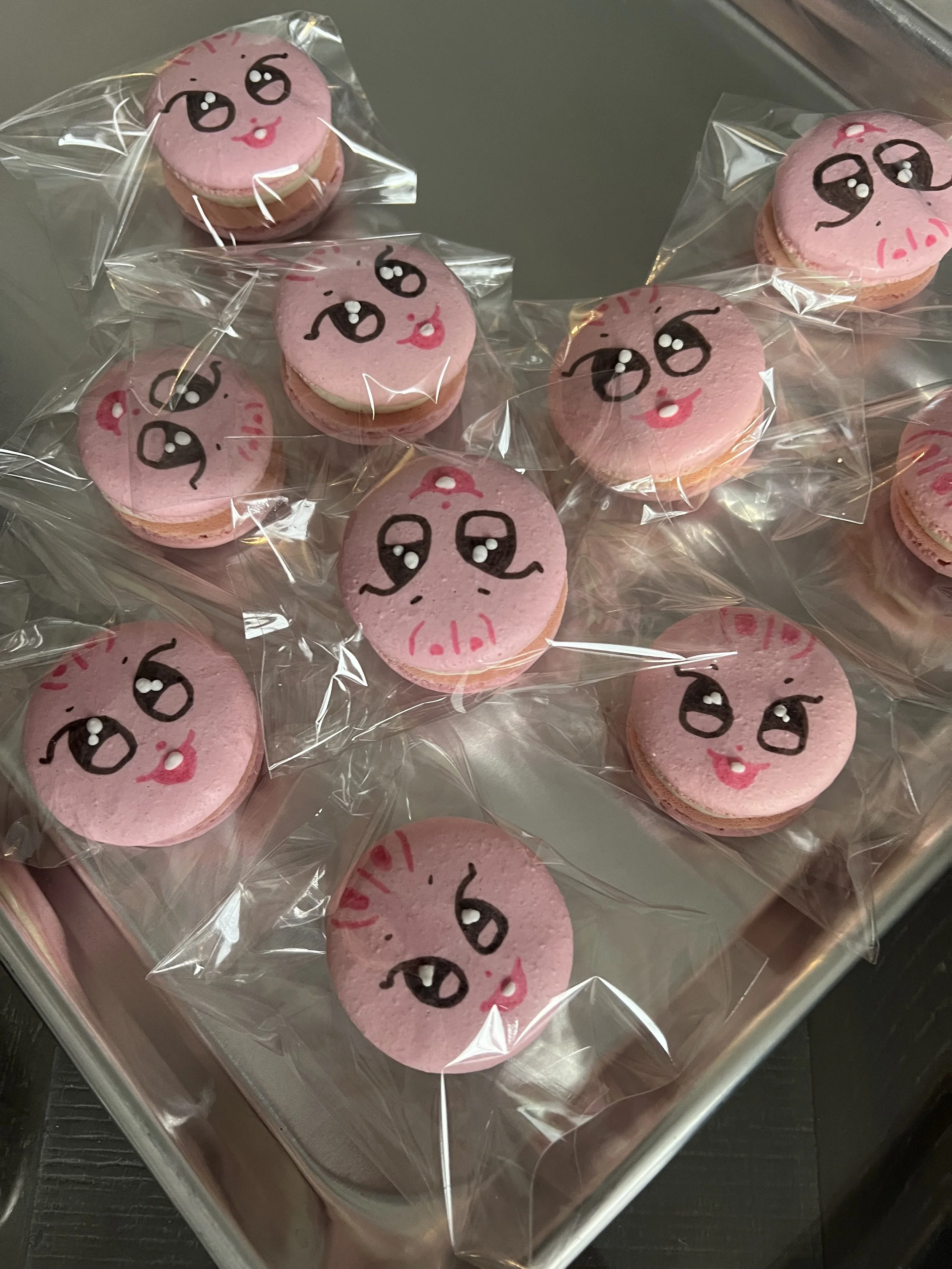 Pink macarons with a cartoon face design, individually wrapped in clear plastic, placed in a metal tray.