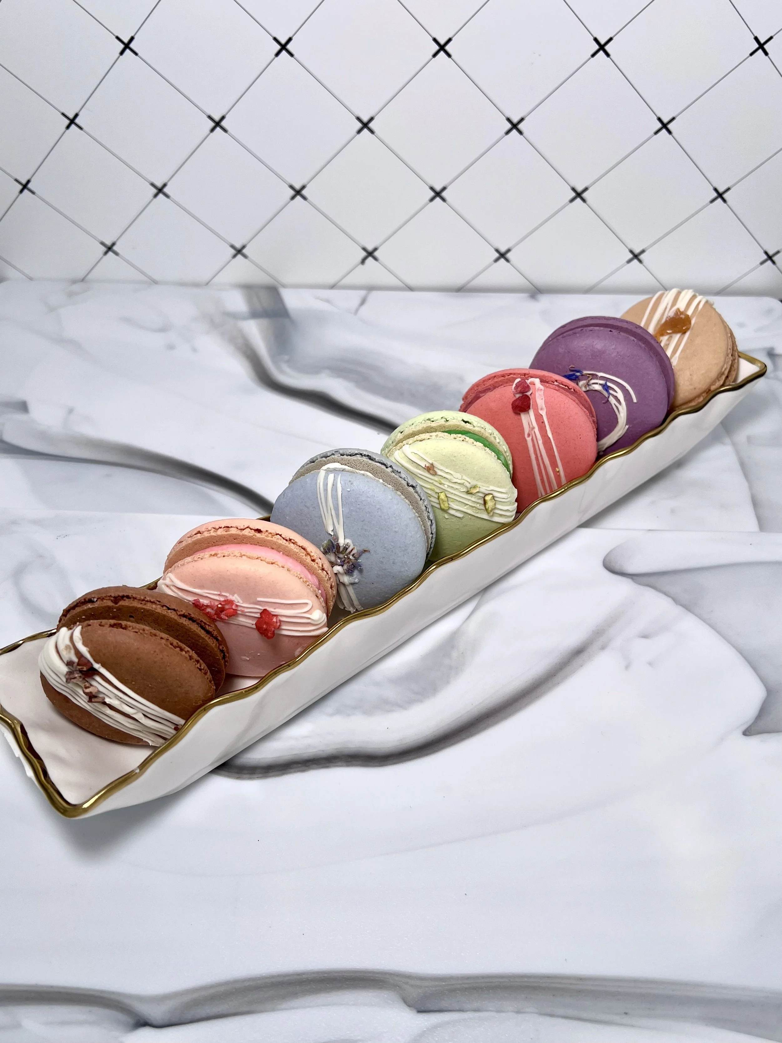A white serving tray with colorful macarons, decorated with white chocolate drizzles and small toppings, resting on a white tablecloth with a crumpled tissue beside it.