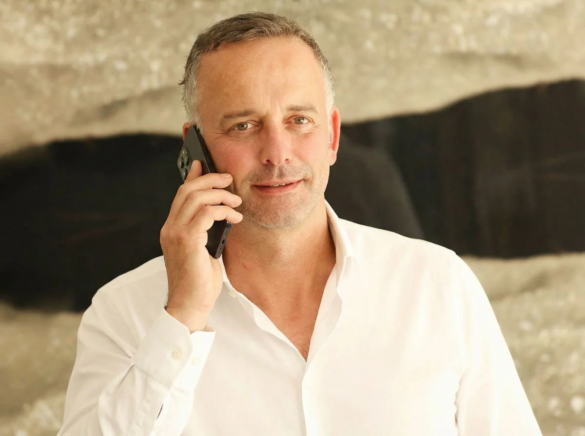 Ein Mann trägt ein weißes Hemd und spricht am Telefon, im Hintergrund eine Wand mit abstrakten Mustern.