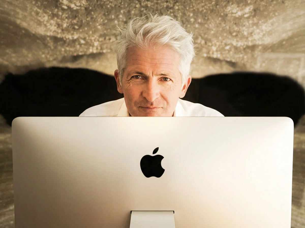 Älterer Mann mit weißen Haaren schaut vor einem Apple-Computer in die Kamera.