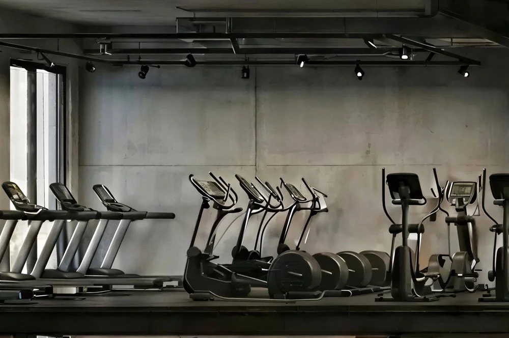 Fitnessraum mit Laufbändern, Ellipsentrainern und Fahrradergometern, in einem modernen, minimalistisch eingerichteten Raum mit Betonwänden und Deckenbeleuchtung.