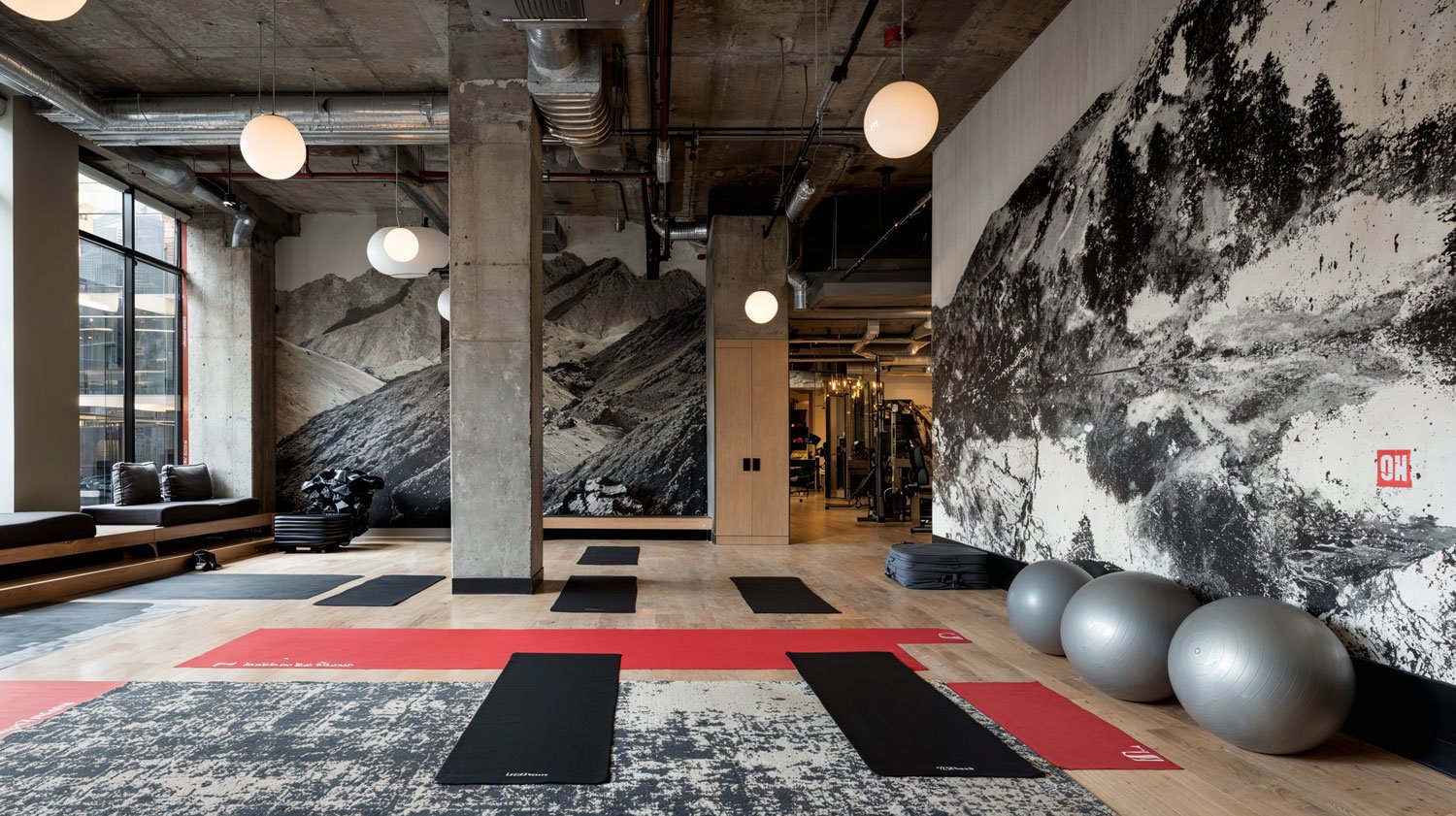 Innenraum eines Fitnessstudios mit schwarzen Yogamatten, Gymnastikbällen, einem weißen Sessel, großen Wandfotos von Berglandschaften und einem Fitnessbereich im Hintergrund.