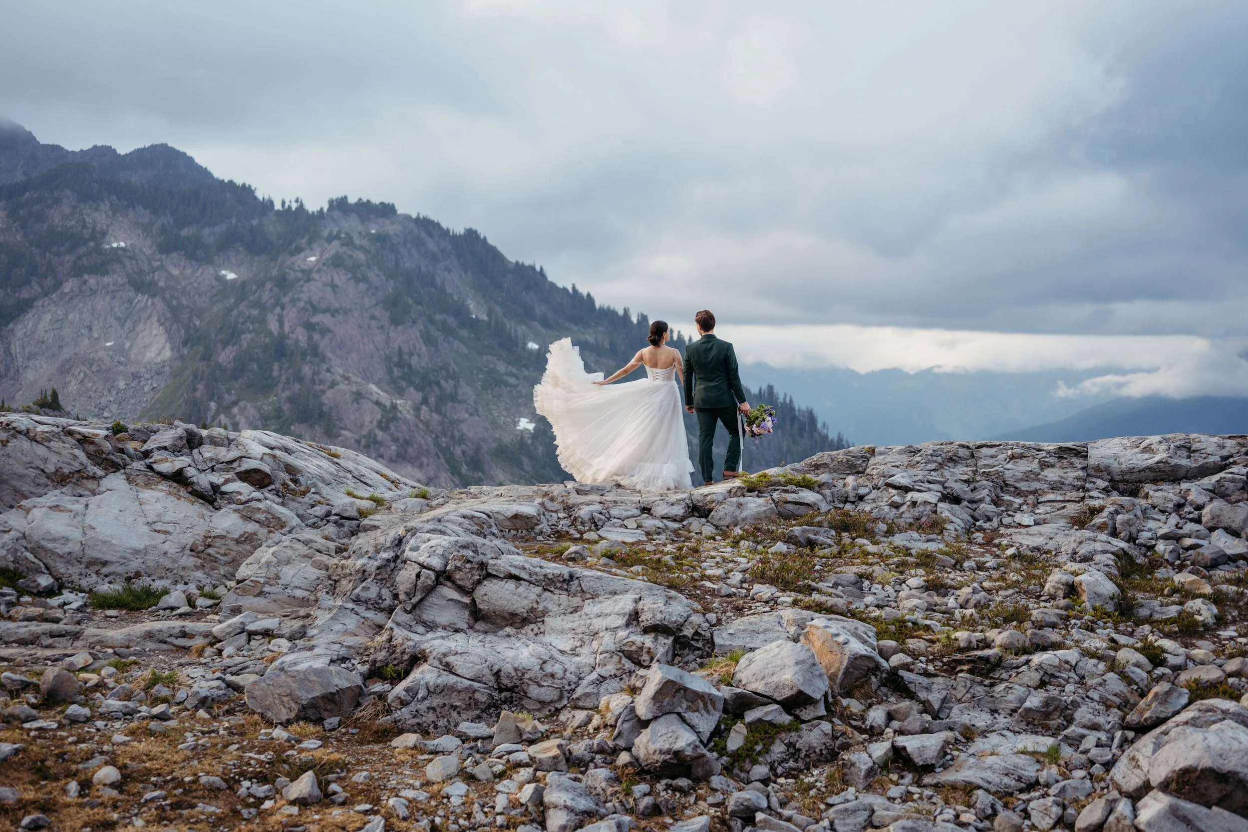A bride and groom standing on rocky terrain outdoors, with a mountainous landscape and cloudy sky in the background.