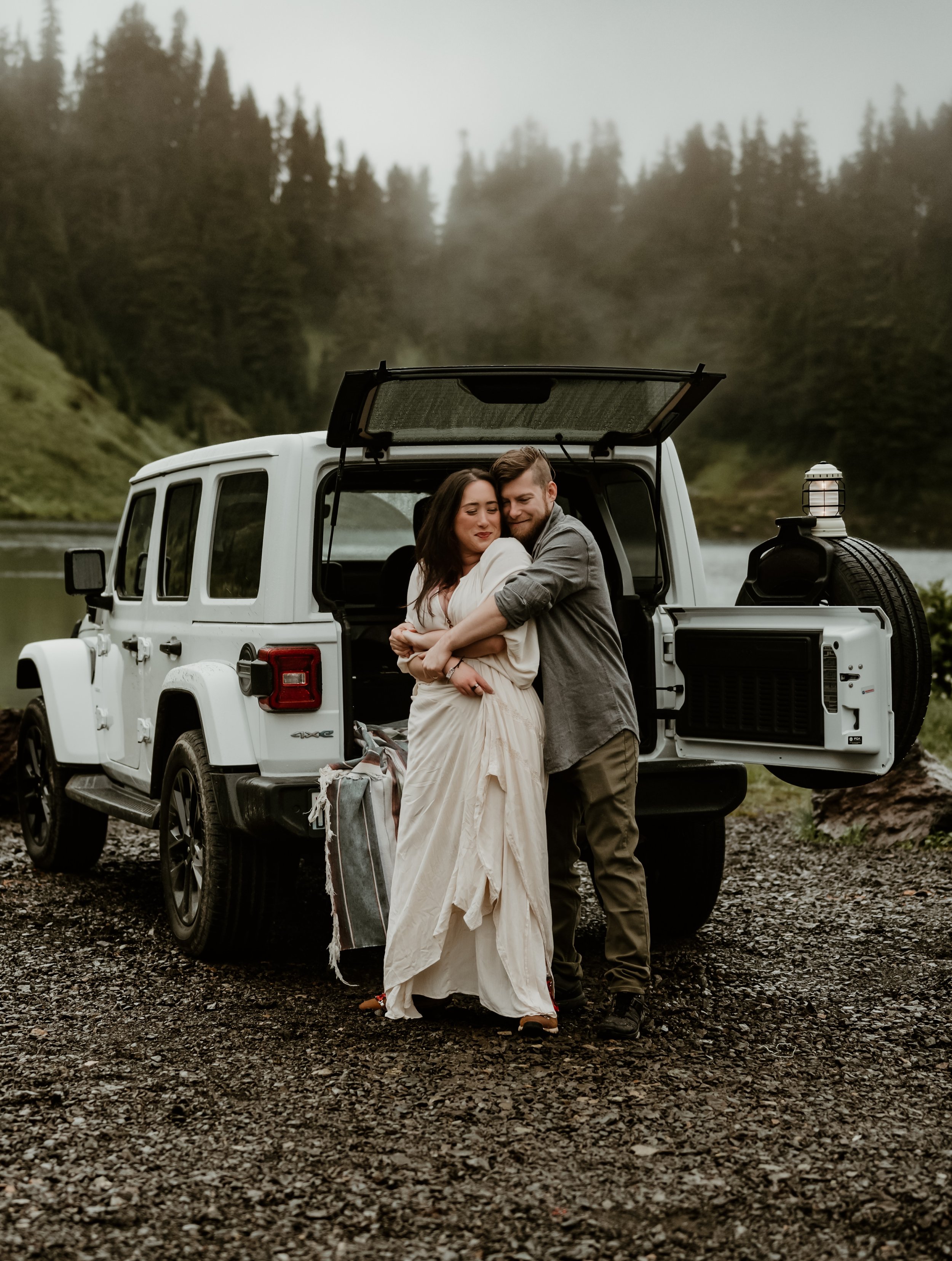 A couple hugging in front of an open back of a white off-road vehicle near a river with a forested mountain landscape in the background. Washington elopement
