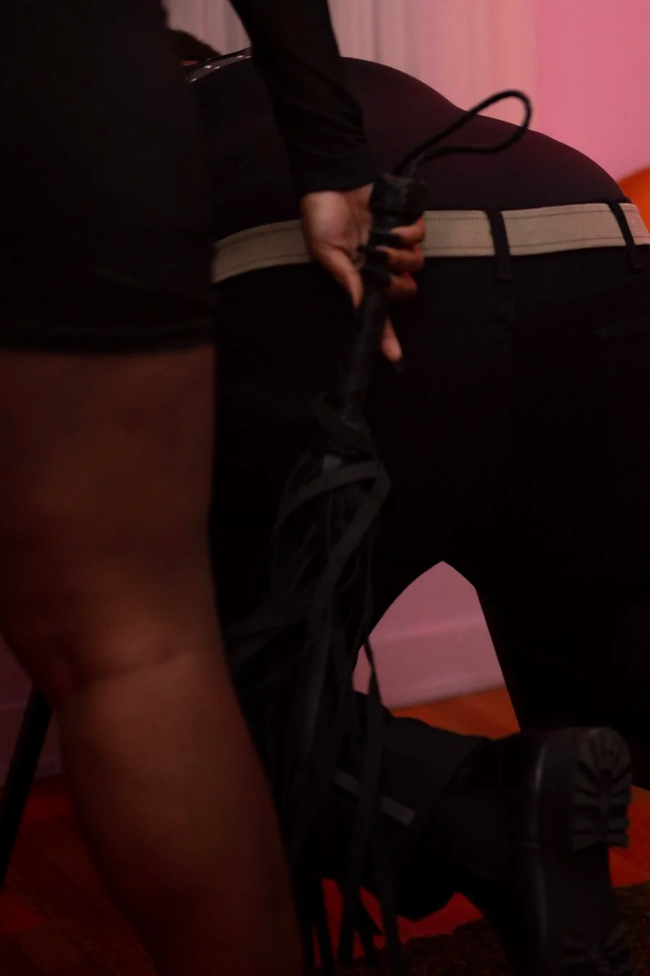 A person in black clothing is kneeling on the floor while another person's hand is holding a black rope or leash.
