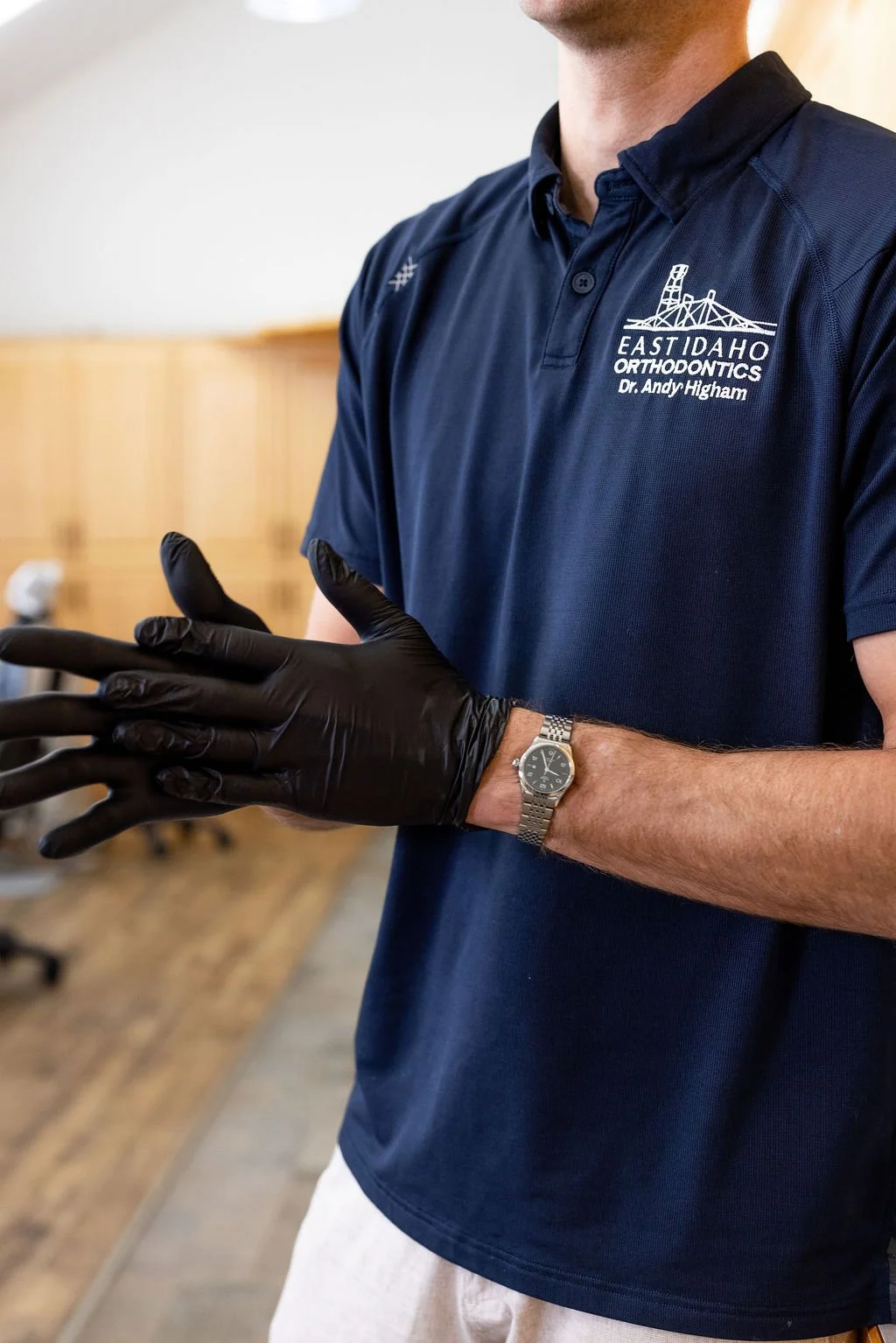 A man wearing a navy blue polo shirt with 'East Idaho Orthodontics Dr. Andy Higham' logo, black gloves, a silver wristwatch, and light-colored pants, standing indoors with a wooden wall in the background.