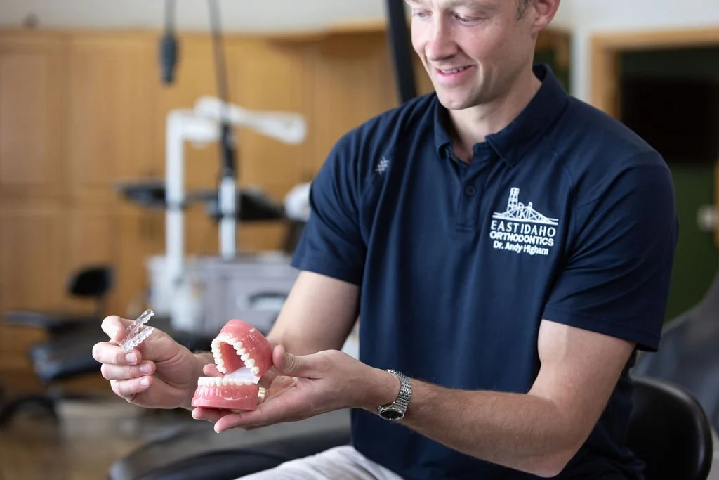 A man in a navy blue polo shirt with East Idaho Orthodontics logo holding a dental model and a clear aligner in an orthodontic office.