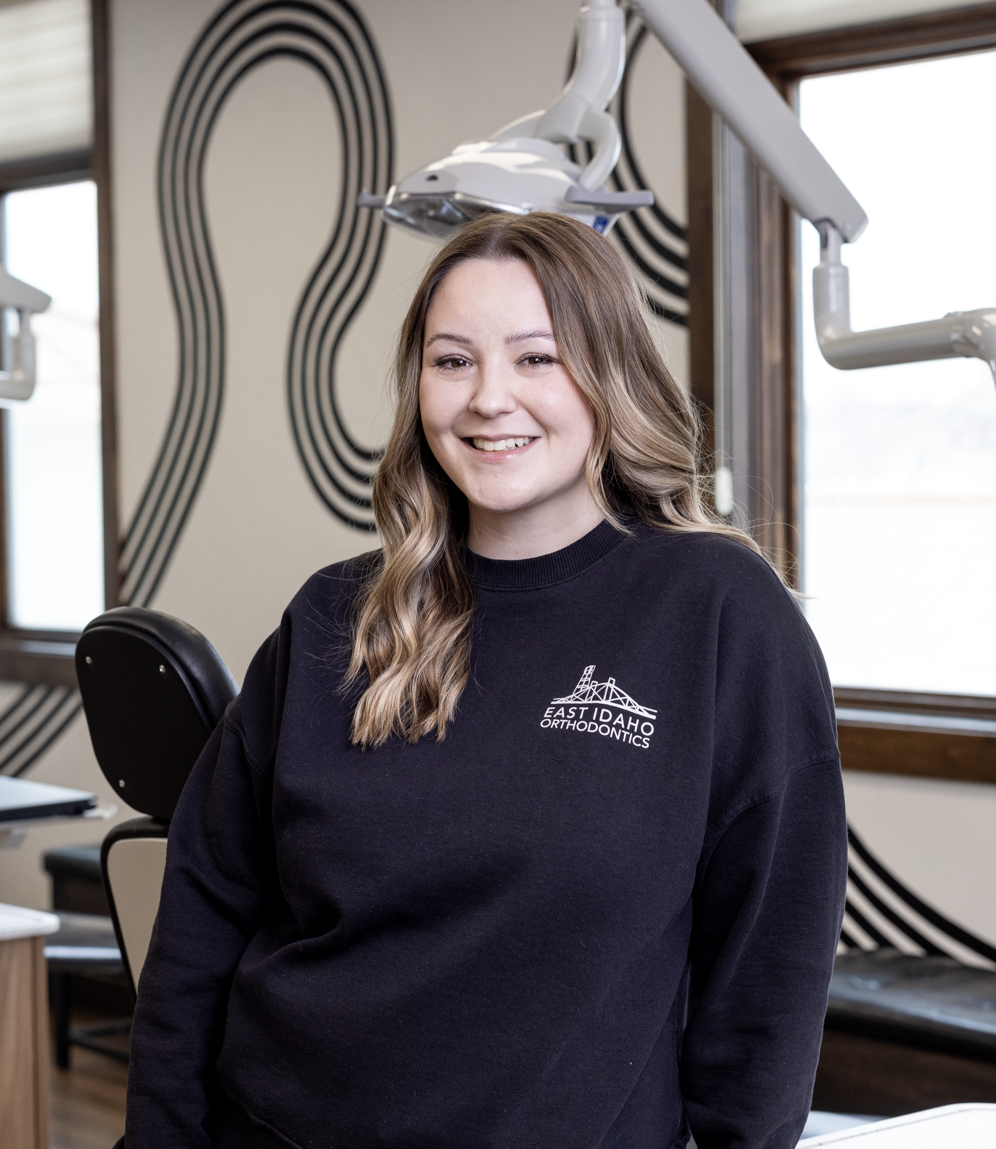A young woman with wavy light brown hair smiling in a dental office, wearing a black sweatshirt with 'East Idaho Orthodontics' logo, dental equipment visible behind her.