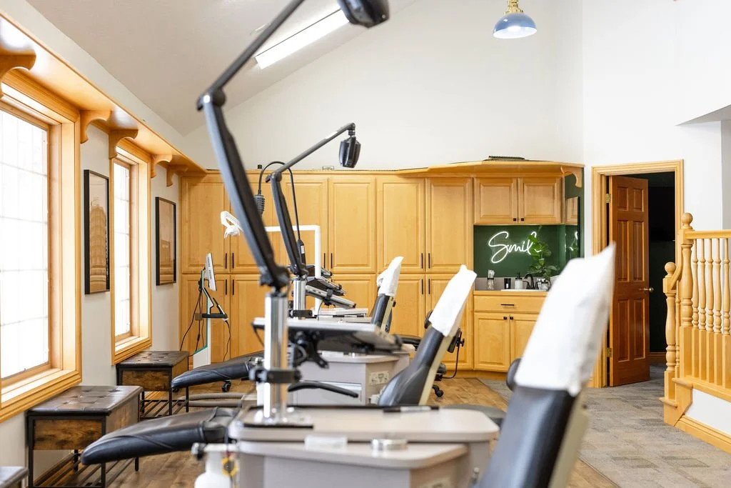 A row of dental chairs in a bright room in Idaho Falls with wooden trim and cabinets, multiple dental lights overhead, and a neon sign that says 'Smile' on the back wall.