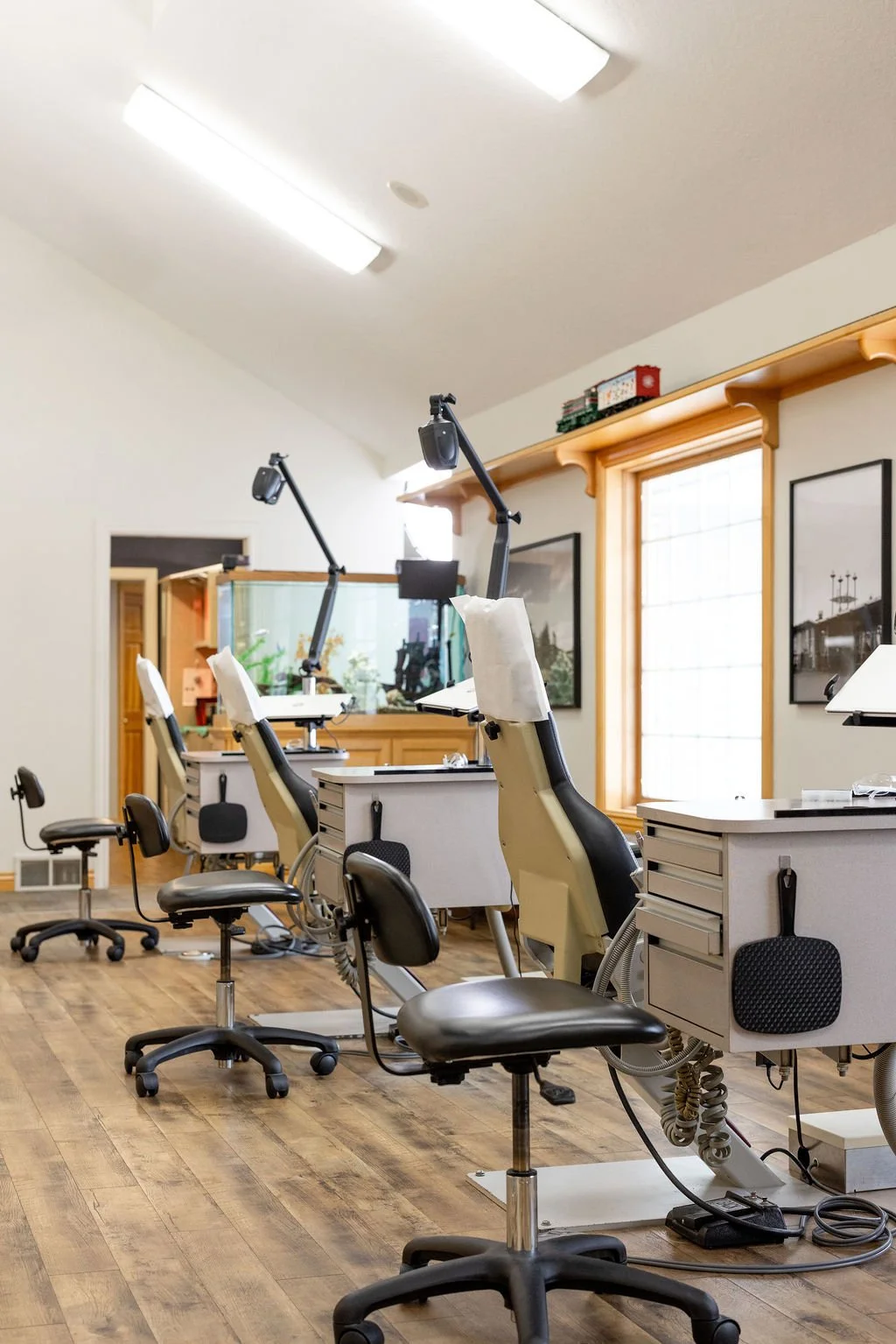 A waiting room with dental chairs and equipment. There are multiple dental chairs with attached lights, along with on wheels stools, in a room with wooden floors and large windows.