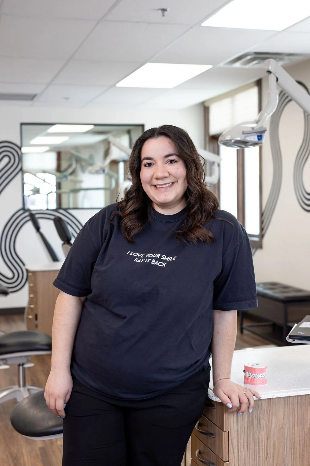A woman smiling in a dental office, wearing a navy blue t-shirt with the text 'I LOVE YOUR SMILE SAY IT BACK,' standing next to a dental model on a counter.