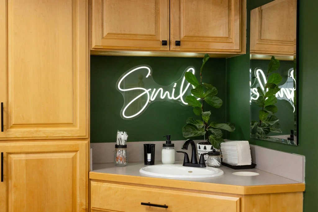 Bathroom with a neon sign that says 'Smile' above the sink, green walls, wooden cabinets, and a mirror reflecting part of the sign.