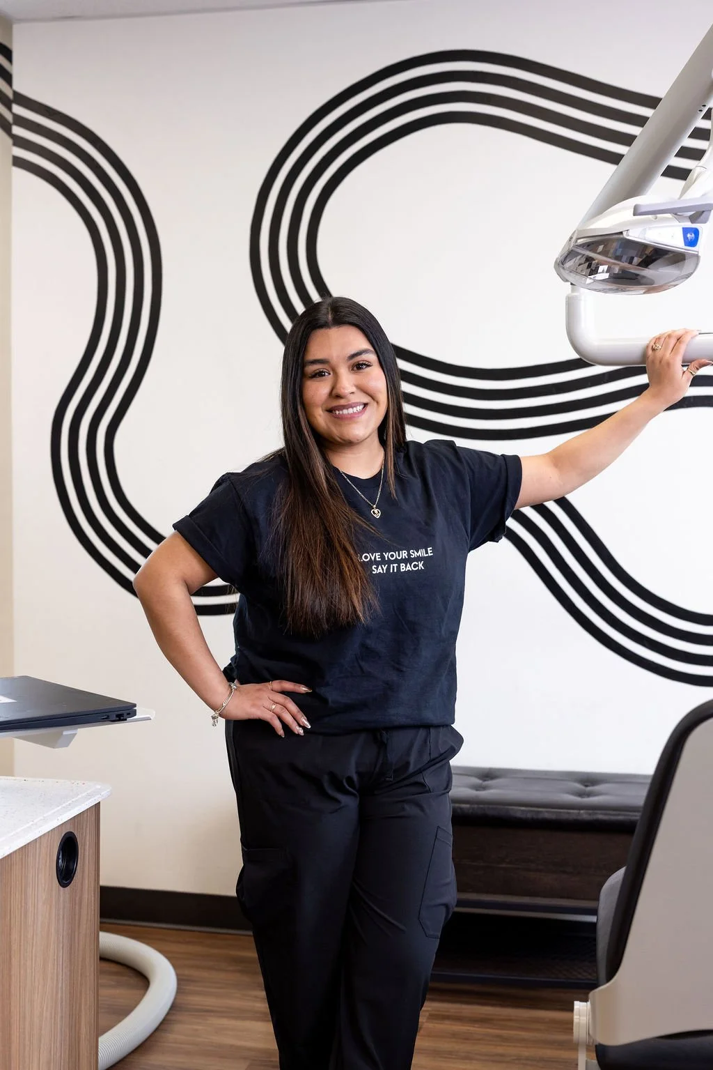 A woman smiling in a dental clinic, wearing a black t-shirt and black pants, standing next to dental equipment with a wall art of black curved lines behind her.