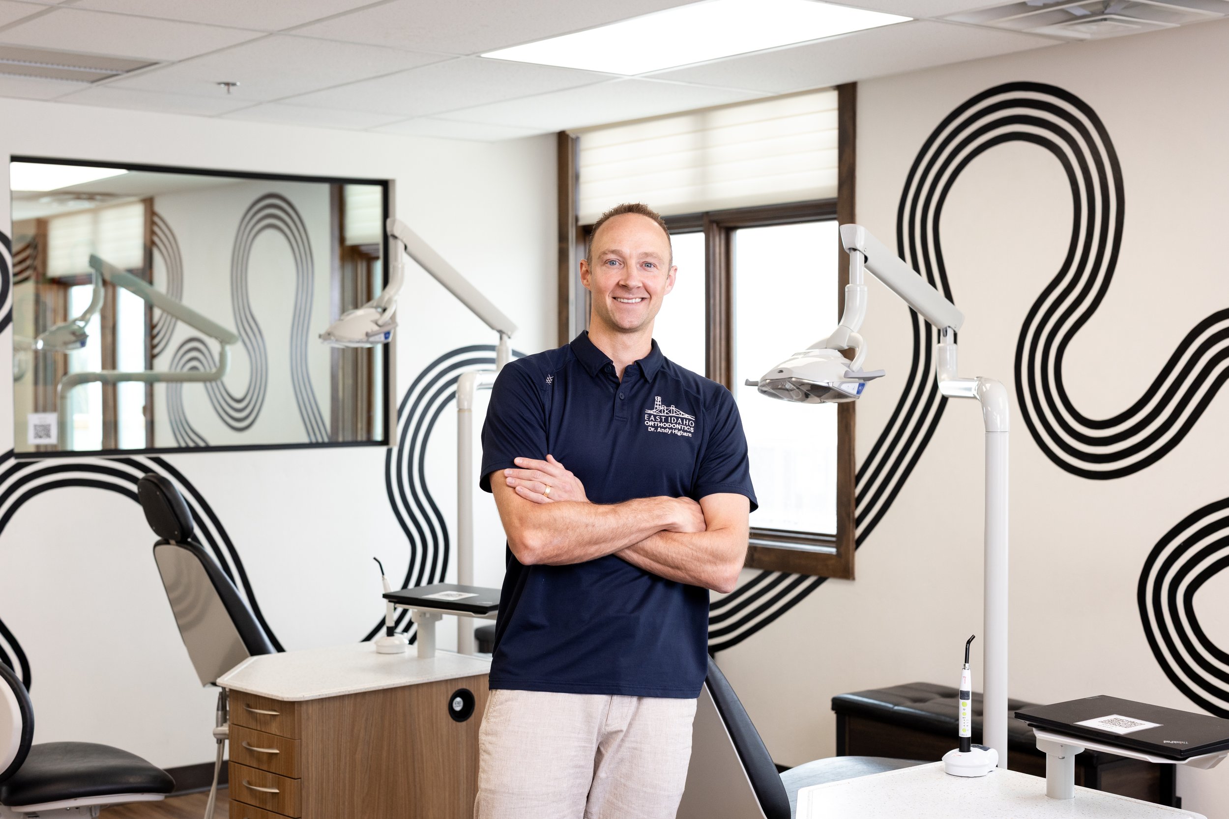 Dr. Higham in a dark blue polo shirt standing in a dental clinic, smiling with arms crossed, with dental equipment and black-and-white abstract wall art in the background.