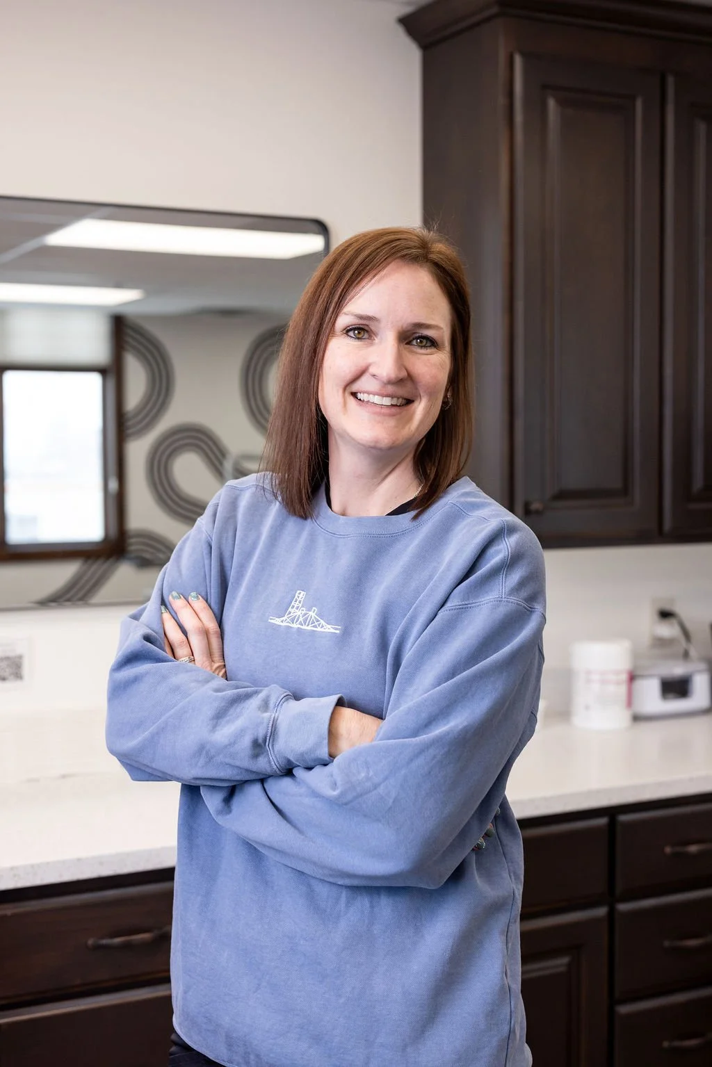 A woman with shoulder-length reddish-brown hair smiling and standing with crossed arms in a kitchen with dark wooden cabinets, a mirror, and a window in the background.